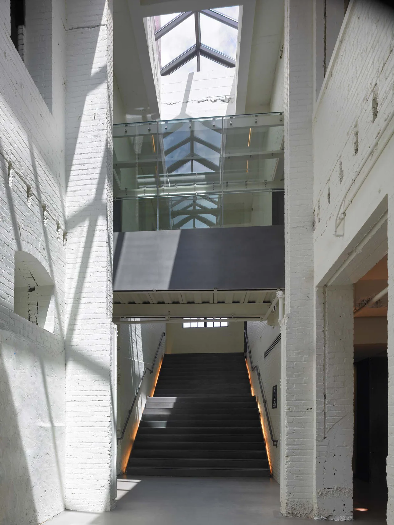 A dark concrete staircase with integrated side lighting is framed by tall white painted brick walls under a large glass skylight.