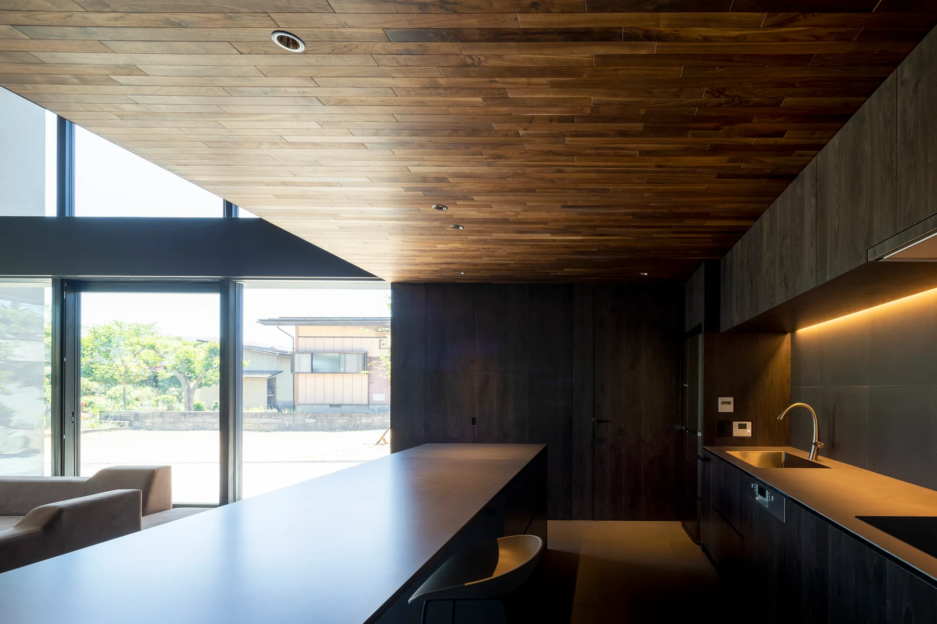 A minimalist kitchen featuring dark flat-panel cabinets, a long dark island, and a warm wood-plank ceiling near large windows.