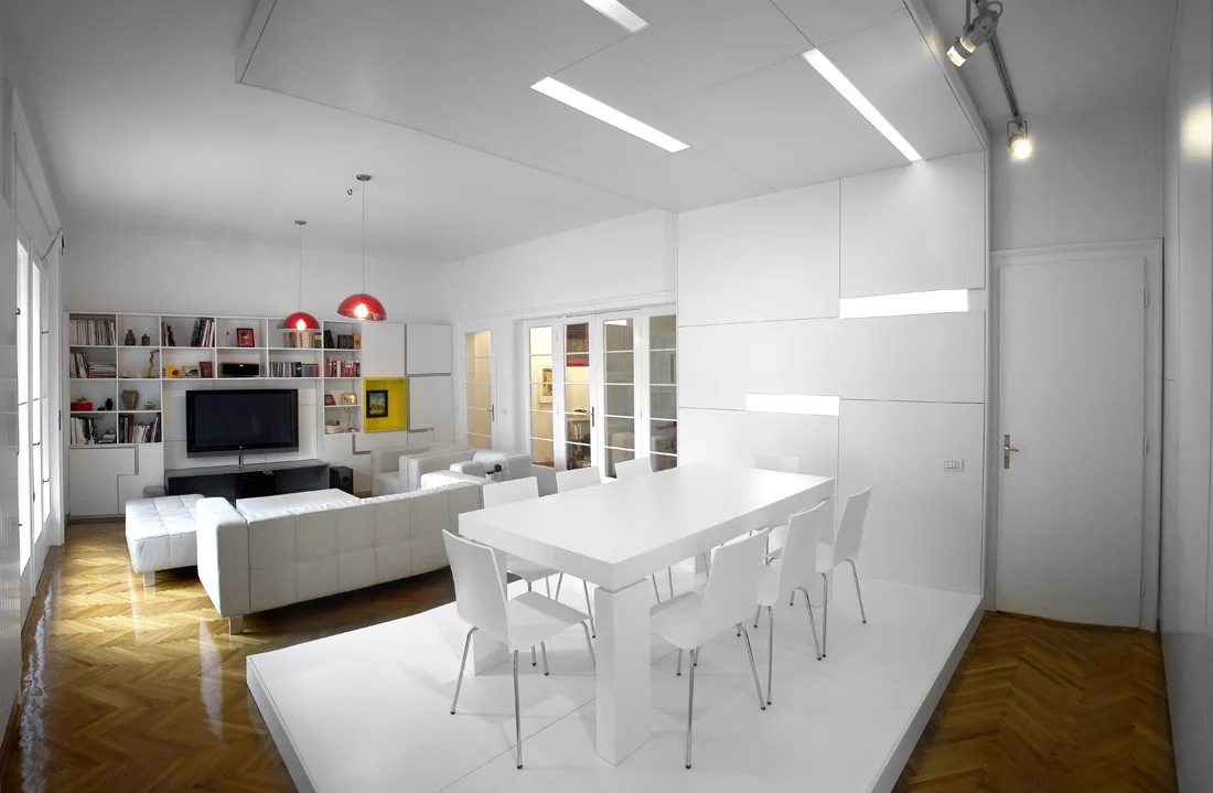 A contemporary open-plan room features a white dining table on a raised platform next to a white leather sofa and polished herringbone wood floors.