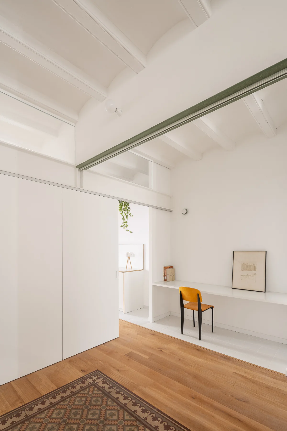 A minimalist workspace features a white floating desk with a single wood chair, set beneath exposed ceiling beams and next to a sliding door.