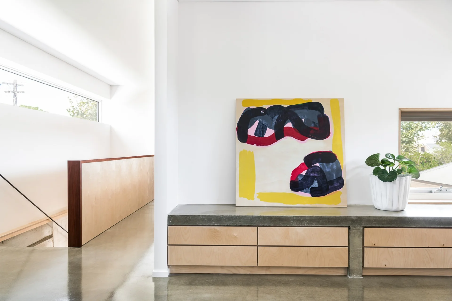 A minimalist hallway landing featuring a low plywood cabinet with a grey concrete top, an abstract painting, and polished concrete floors.