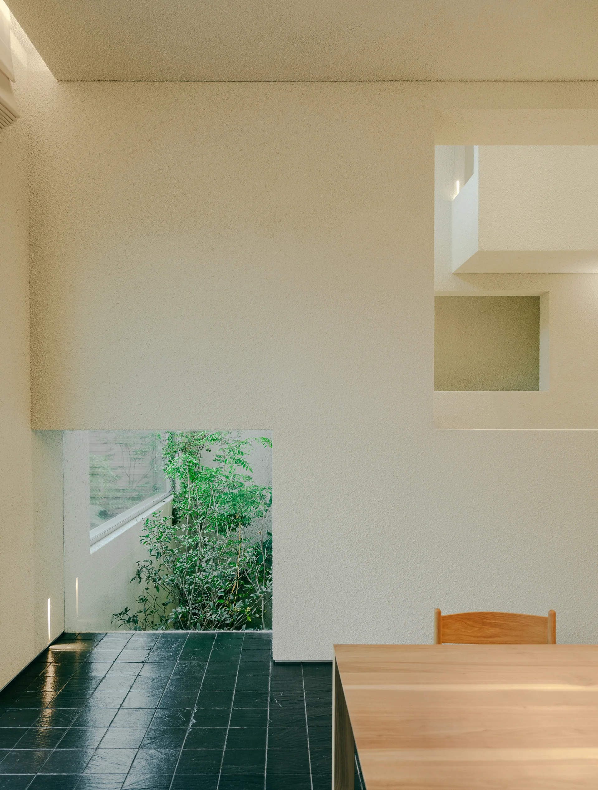 A minimalist dining area features a large textured plaster wall with geometric cutouts, a dark tiled floor, and a wooden table in the foreground.