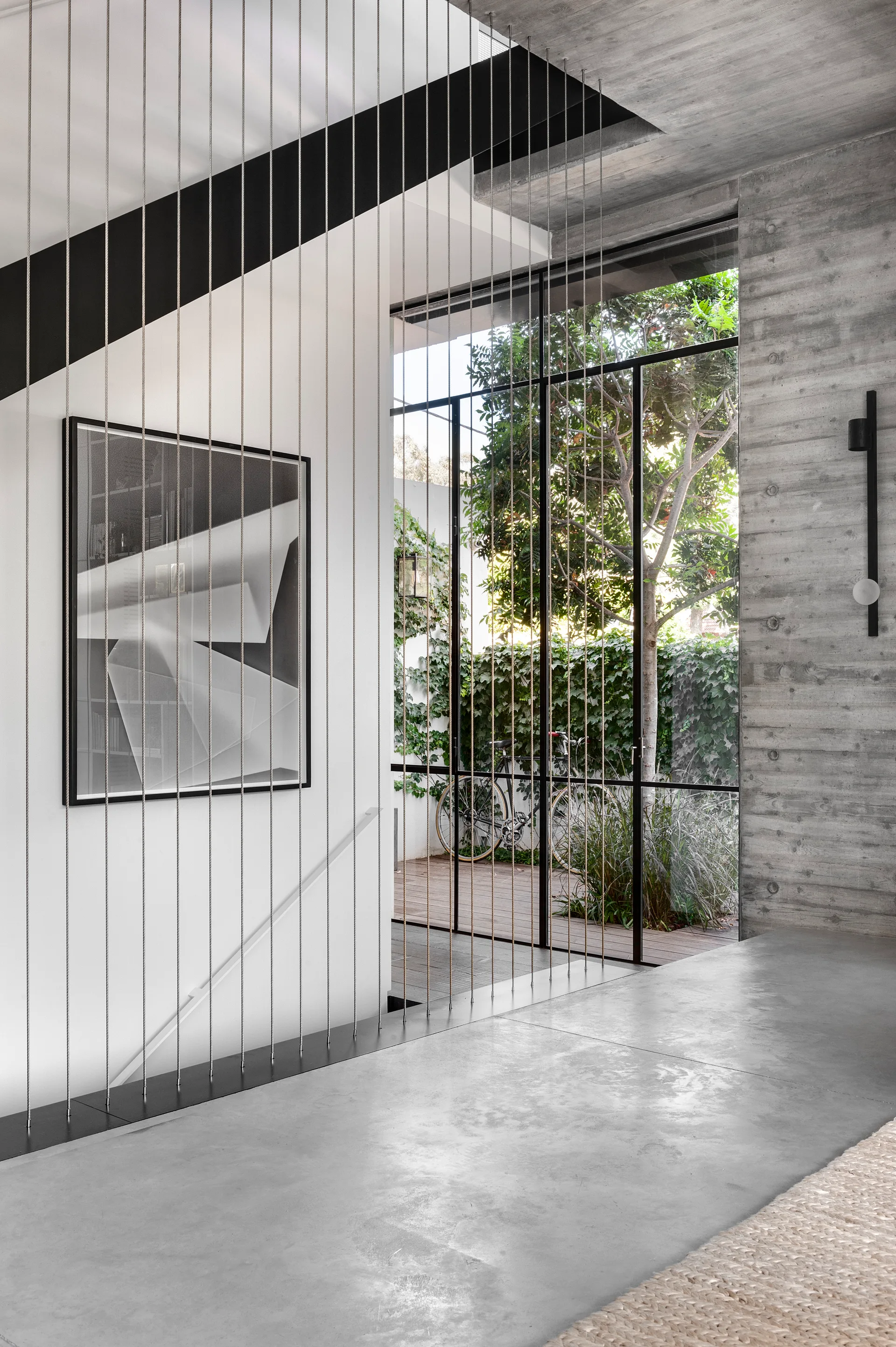 A modern entryway featuring vertical tensioned cable stair railing, board-formed concrete walls, and a glass door opening to a garden.