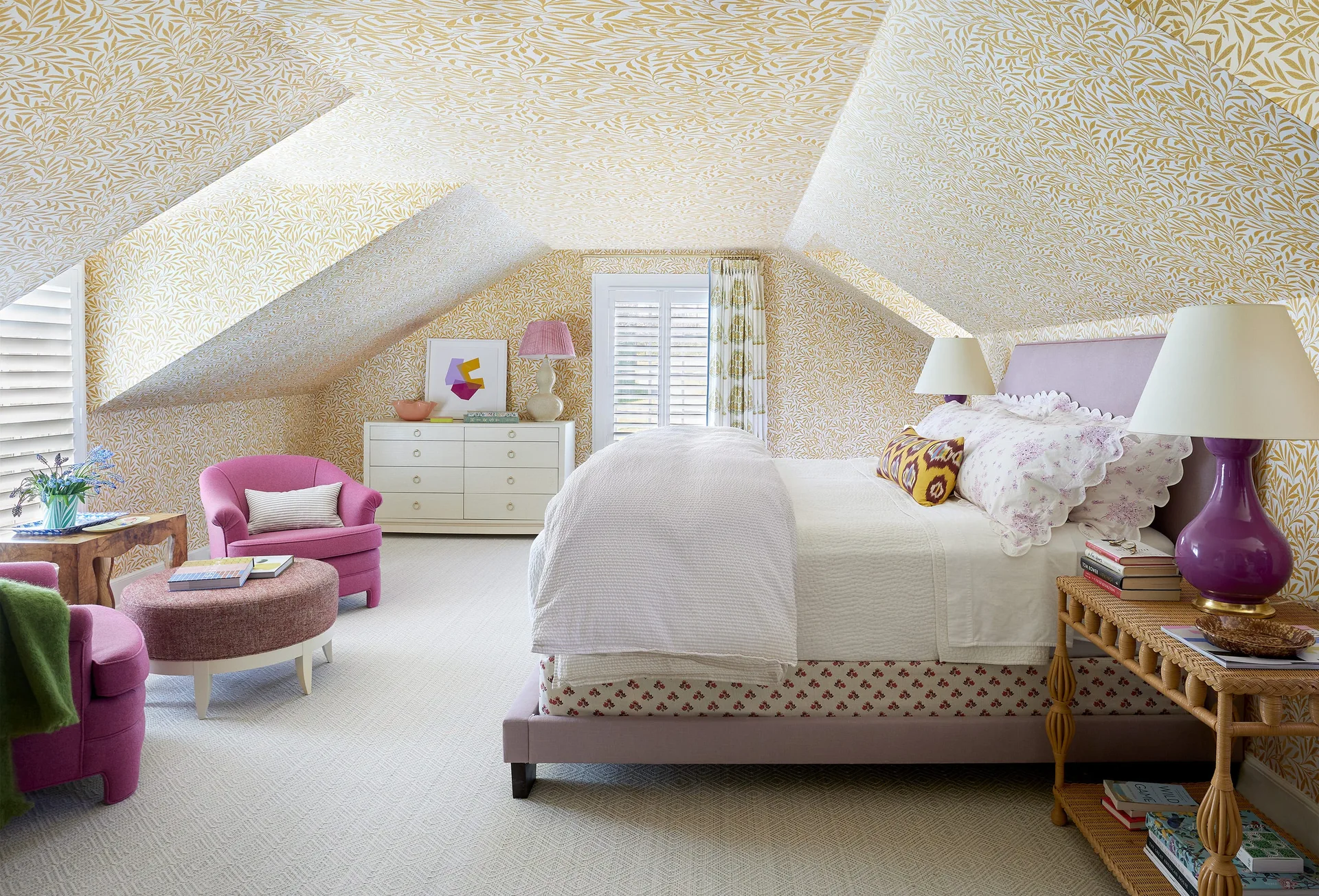 An attic bedroom features yellow patterned wallpaper covering the sloped ceilings, a lavender upholstered bed, pink armchairs, and a rattan nightstand.