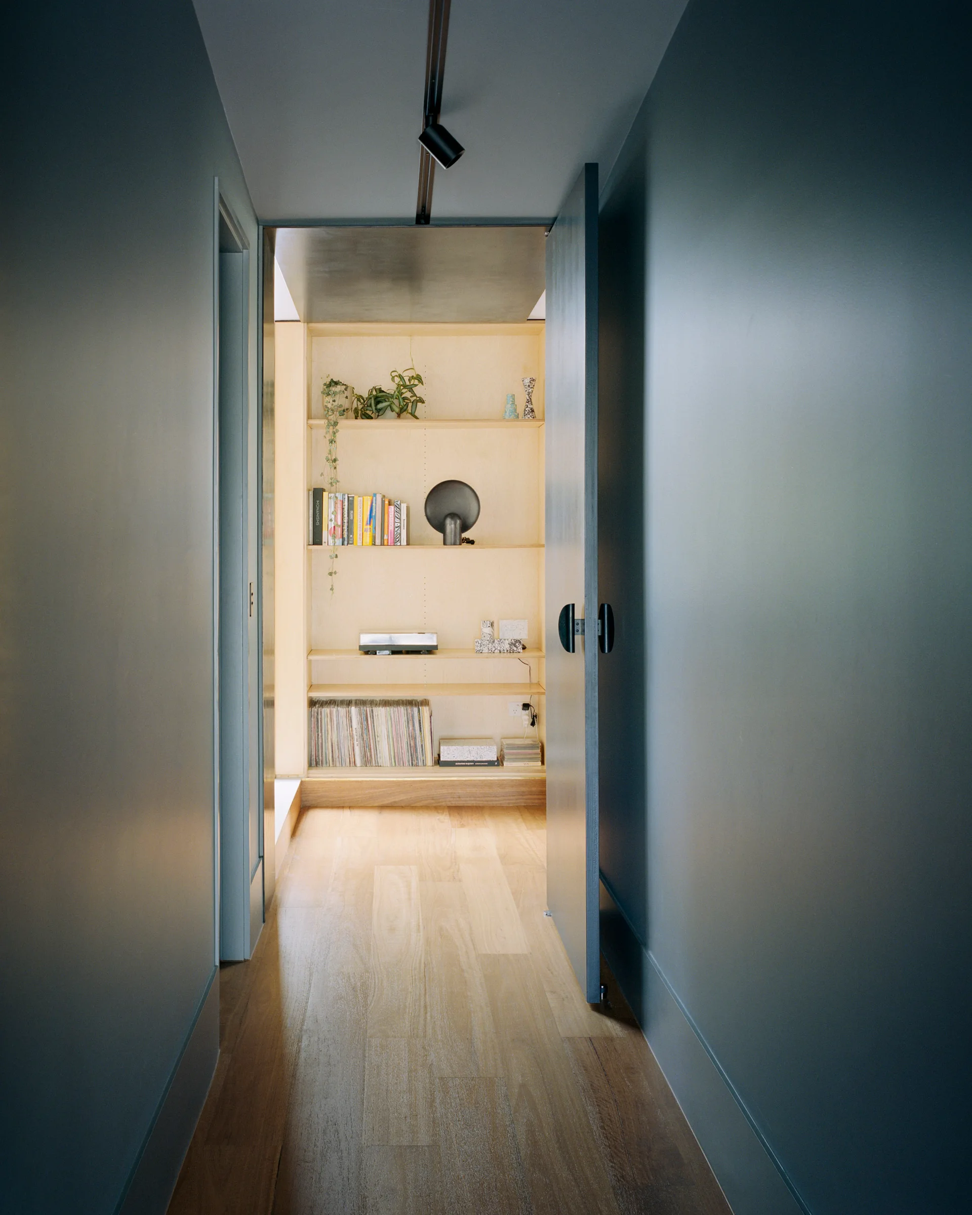 A narrow hallway with dark blue walls leads to a brightly lit wooden alcove containing built-in shelving filled with books and vinyl records.