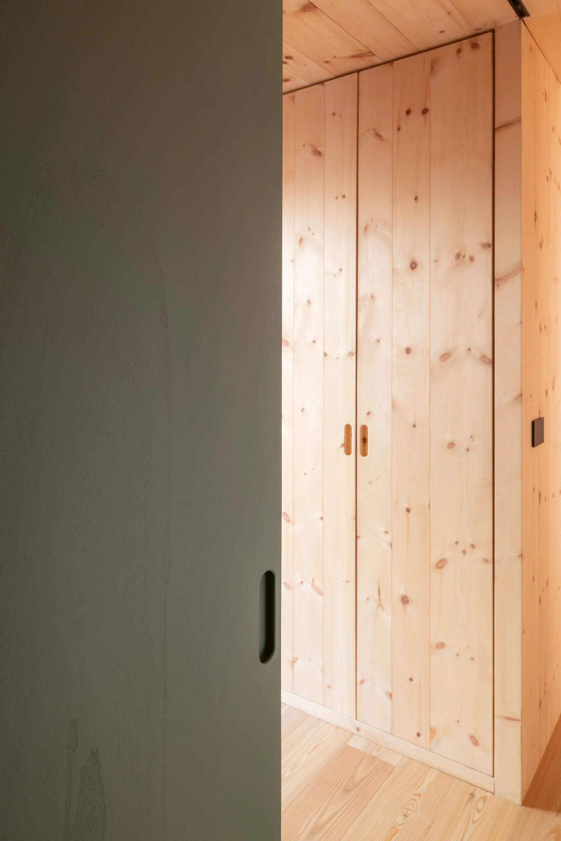 A view through a grey sliding door reveals a custom wardrobe made of light knotty wood planks with recessed circular pull handles.