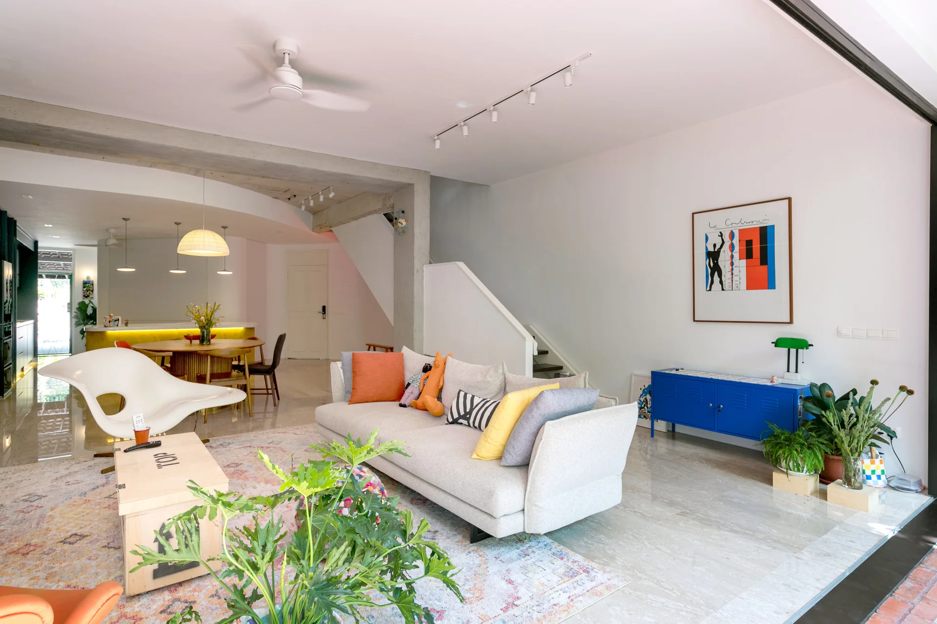 An open-plan living room features a light grey sofa, a blue metal locker cabinet, an exposed concrete beam, and a polished tile floor.