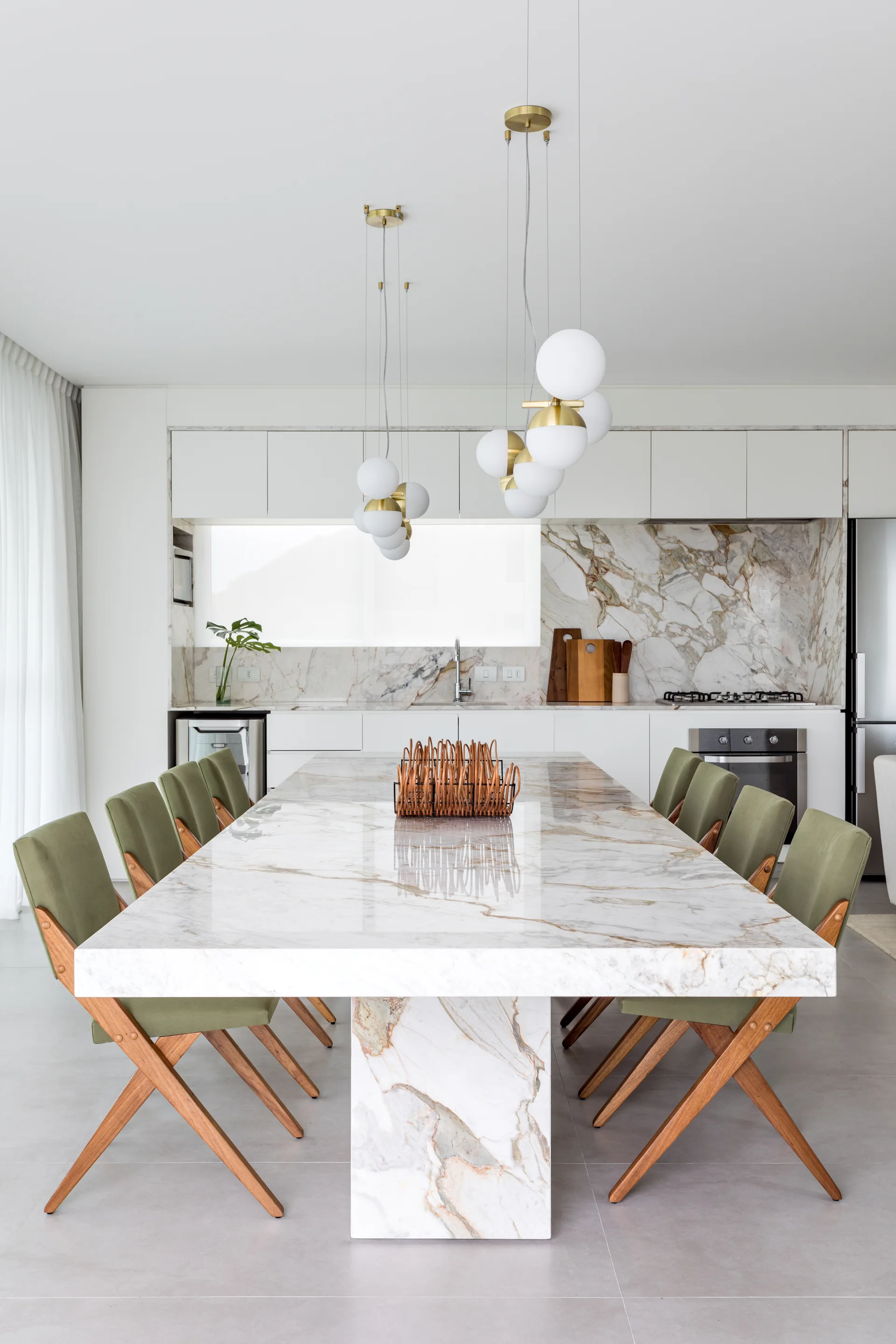 A large veined white marble dining table is surrounded by sage green chairs under globe pendant lights, with a kitchen in the background.