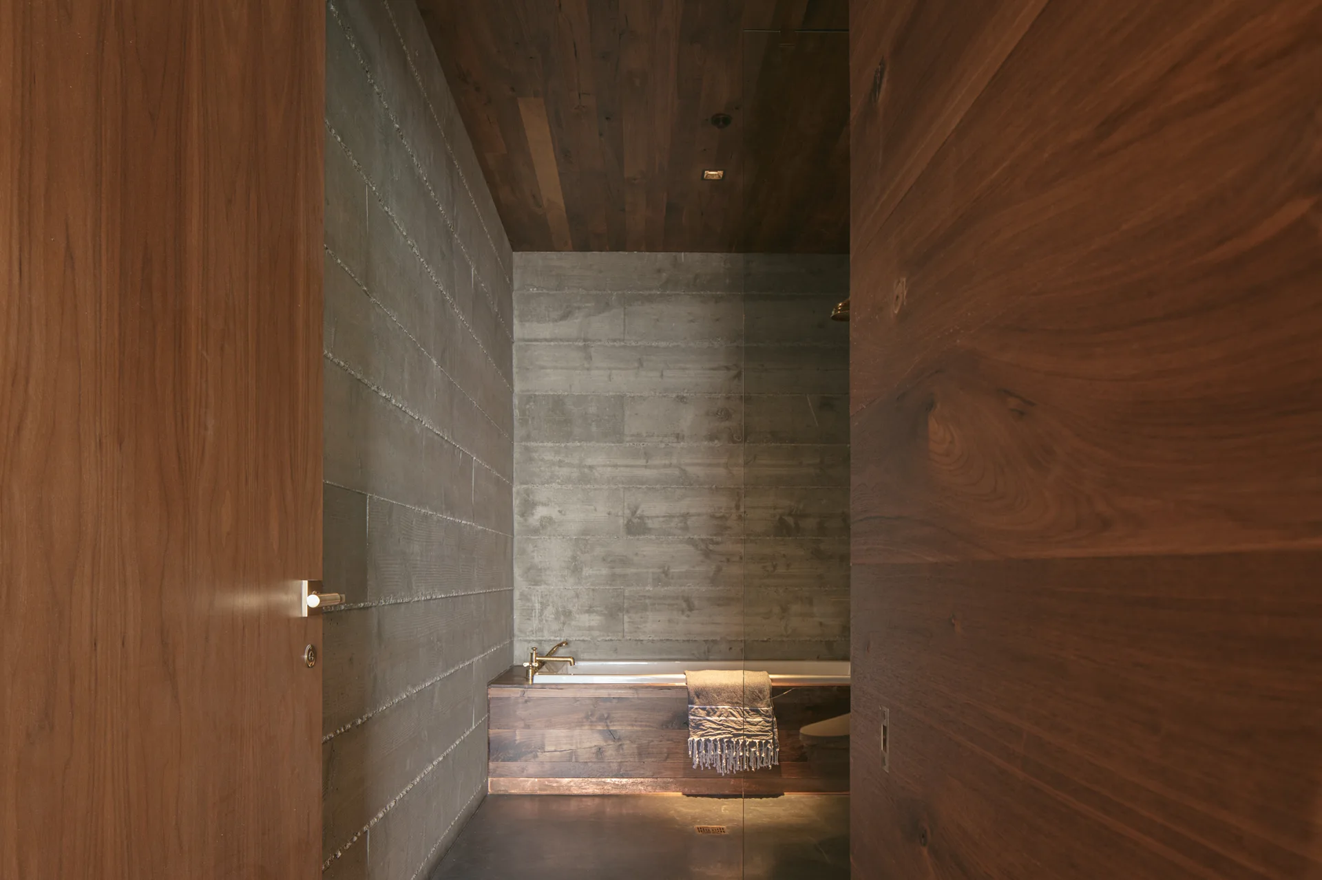 A minimalist bathroom viewed through a wooden doorway, featuring horizontal concrete walls, a dark wood ceiling, and a bathtub with brass hardware.