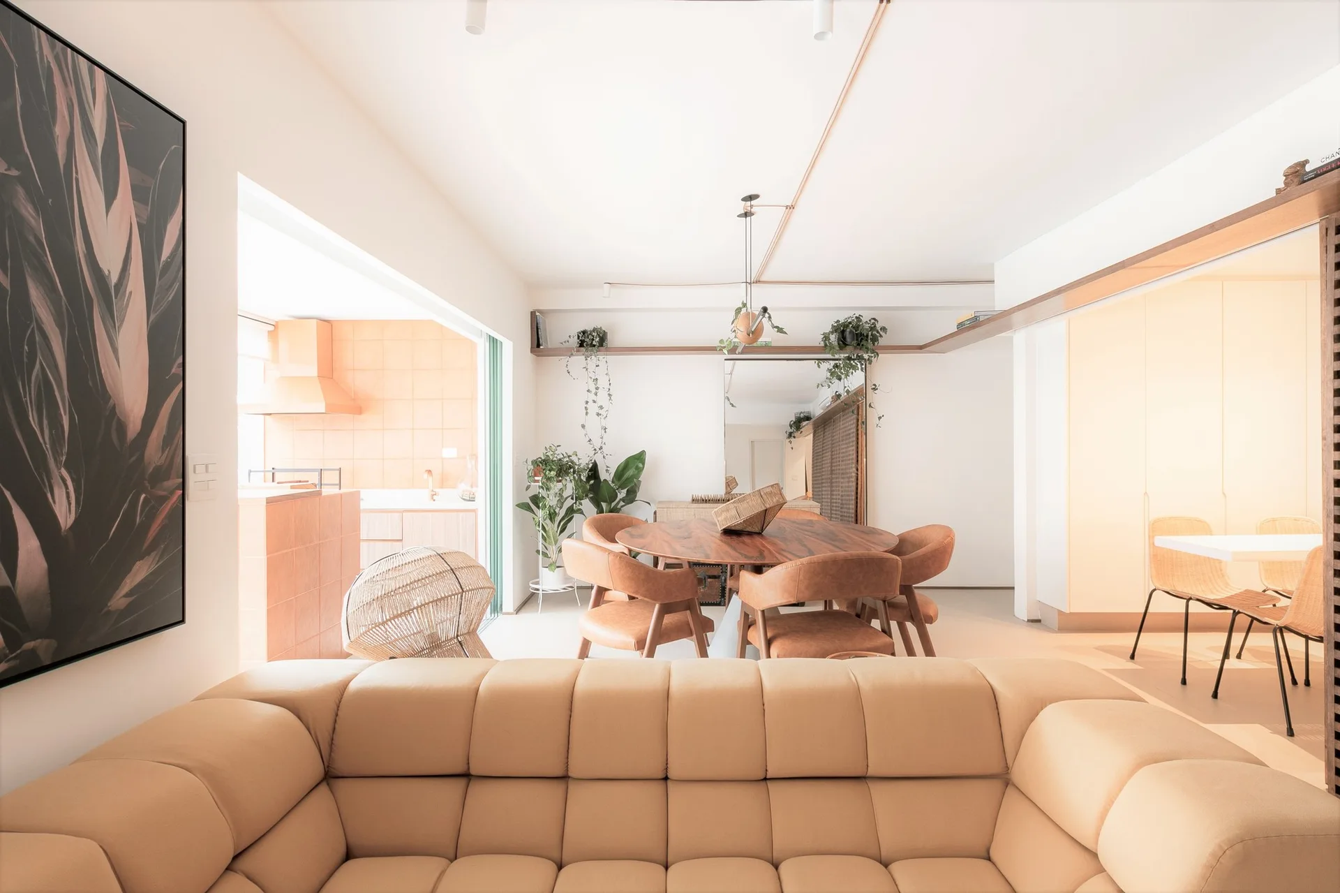 An open living room with a tufted tan sofa, a round wood dining table with leather chairs, and a terracotta tiled kitchen visible in the background.