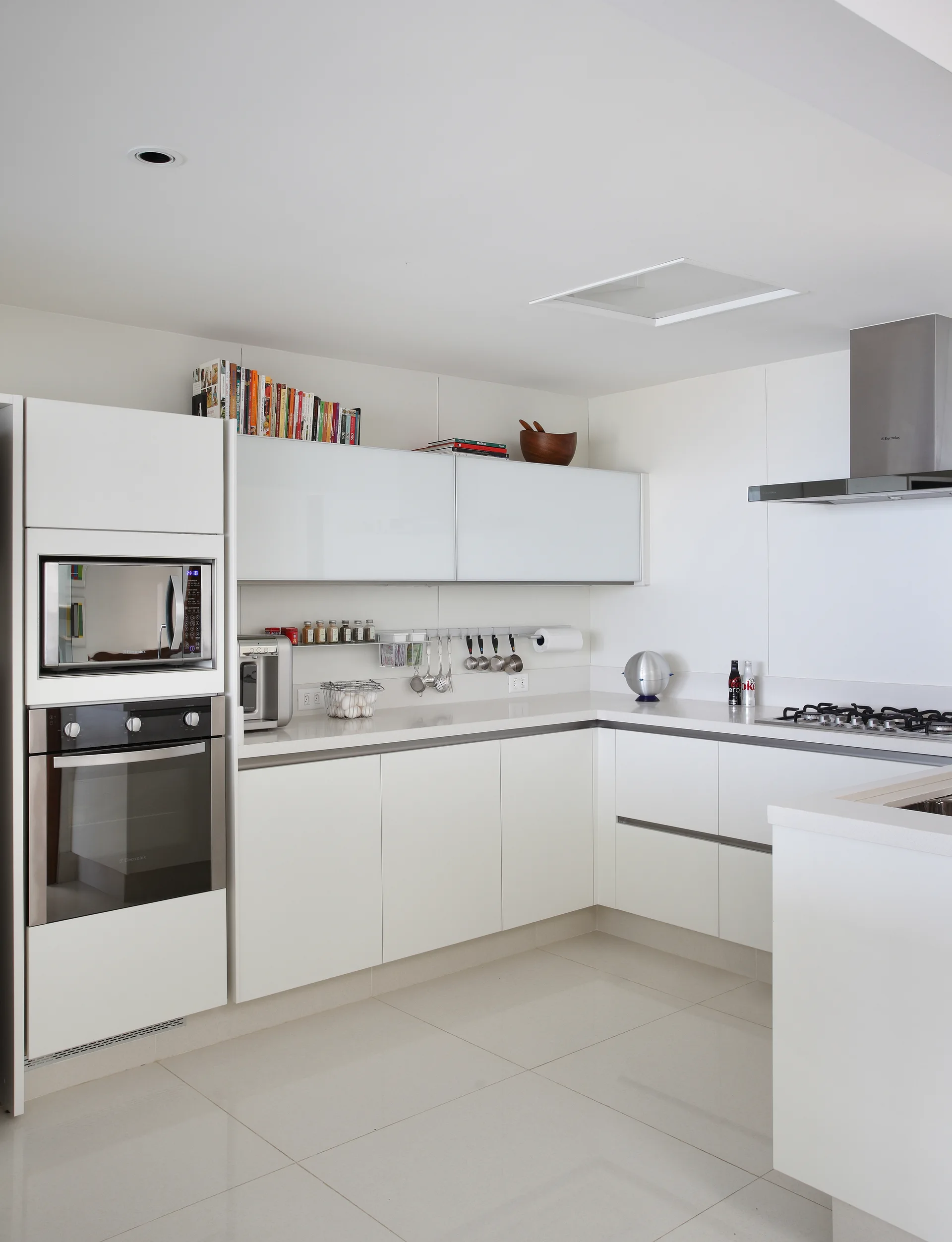 A minimalist white kitchen featuring glossy slab-front cabinets, a built-in oven stack, stainless steel range hood, and large light-colored floor tiles.