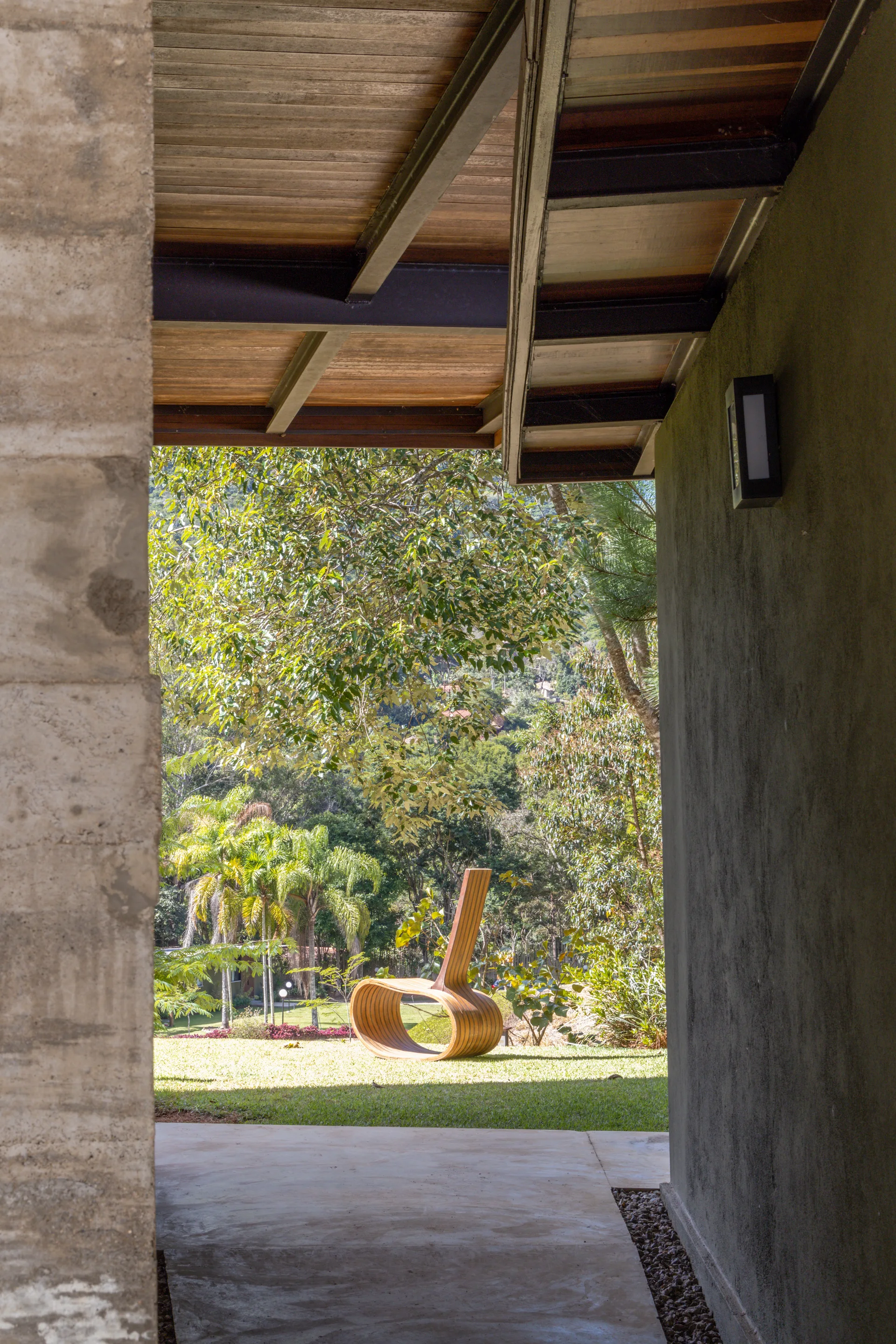 A view from a covered concrete patio looking out to a grassy garden with a sculptural wooden lounge chair.