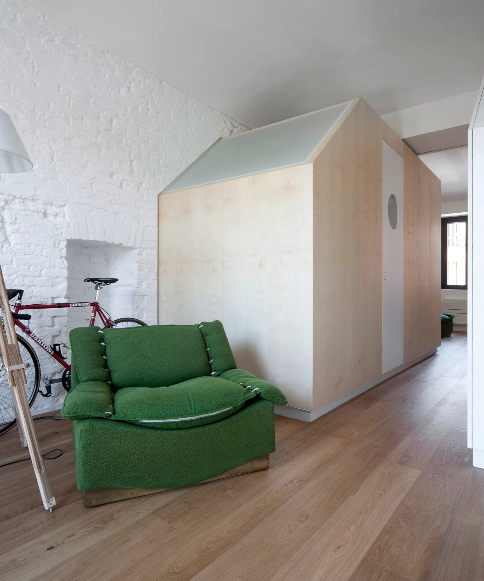 A green armchair sits near a white painted brick wall and a custom house-shaped plywood room pod.