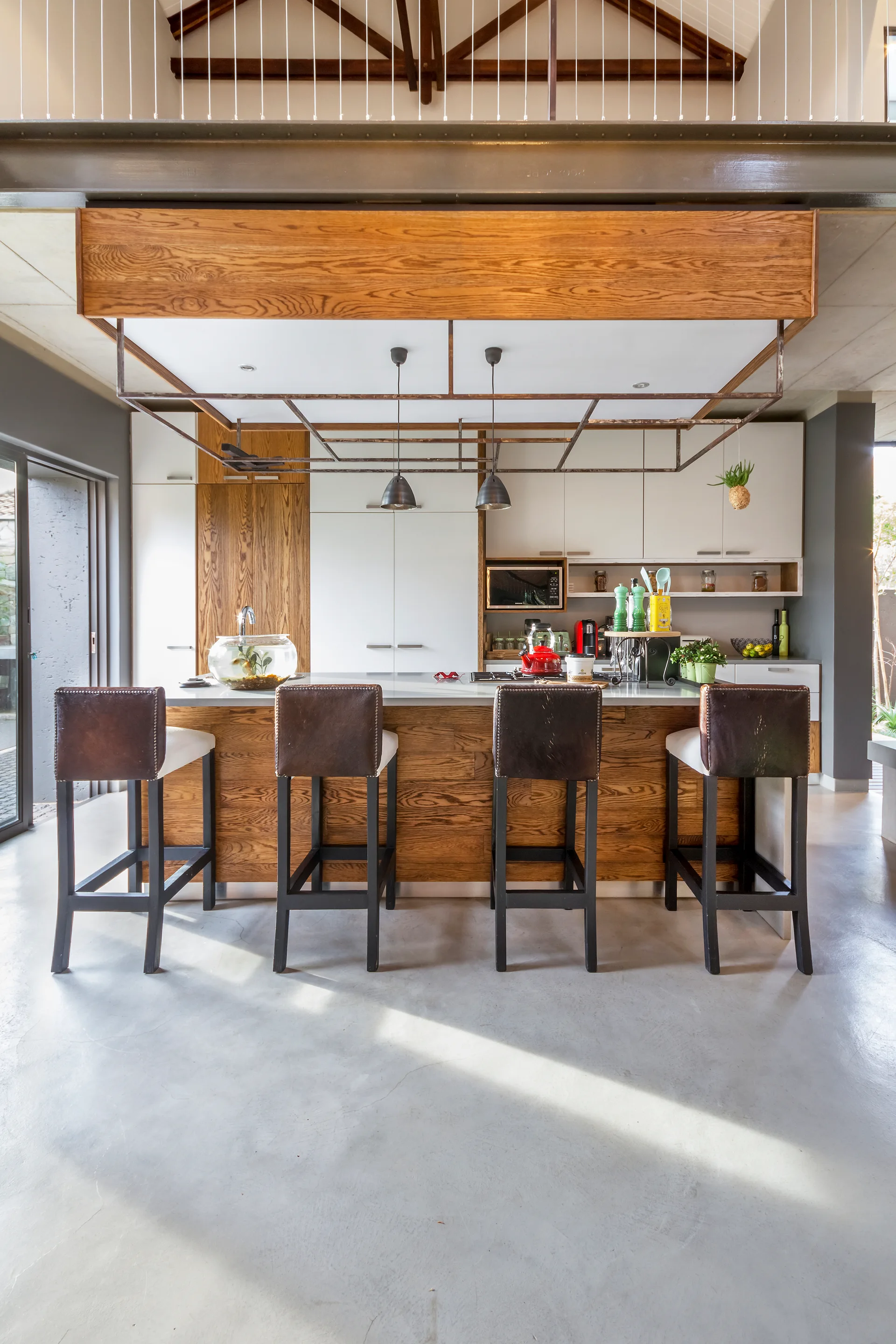 Four leather bar stools sit at a wood-paneled kitchen island beneath a suspended metal frame and an exposed wood truss ceiling.
