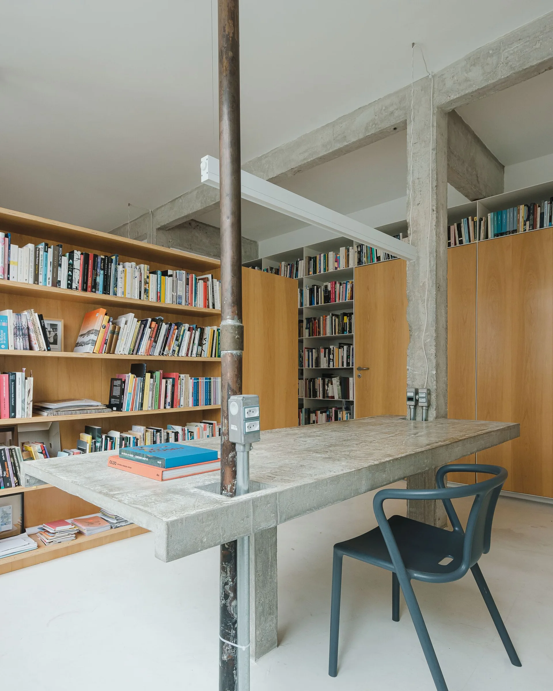 A concrete desk with an integrated metal conduit pipe stands in an industrial office next to floor-to-ceiling wooden bookshelves.