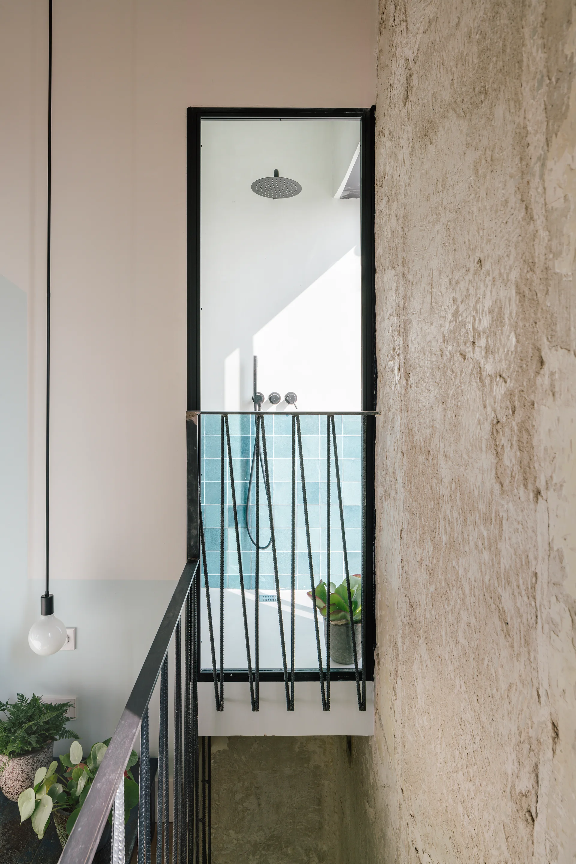 A view from a staircase landing through a black-framed window into a shower with teal tiles, next to a rough plaster wall and black railing.