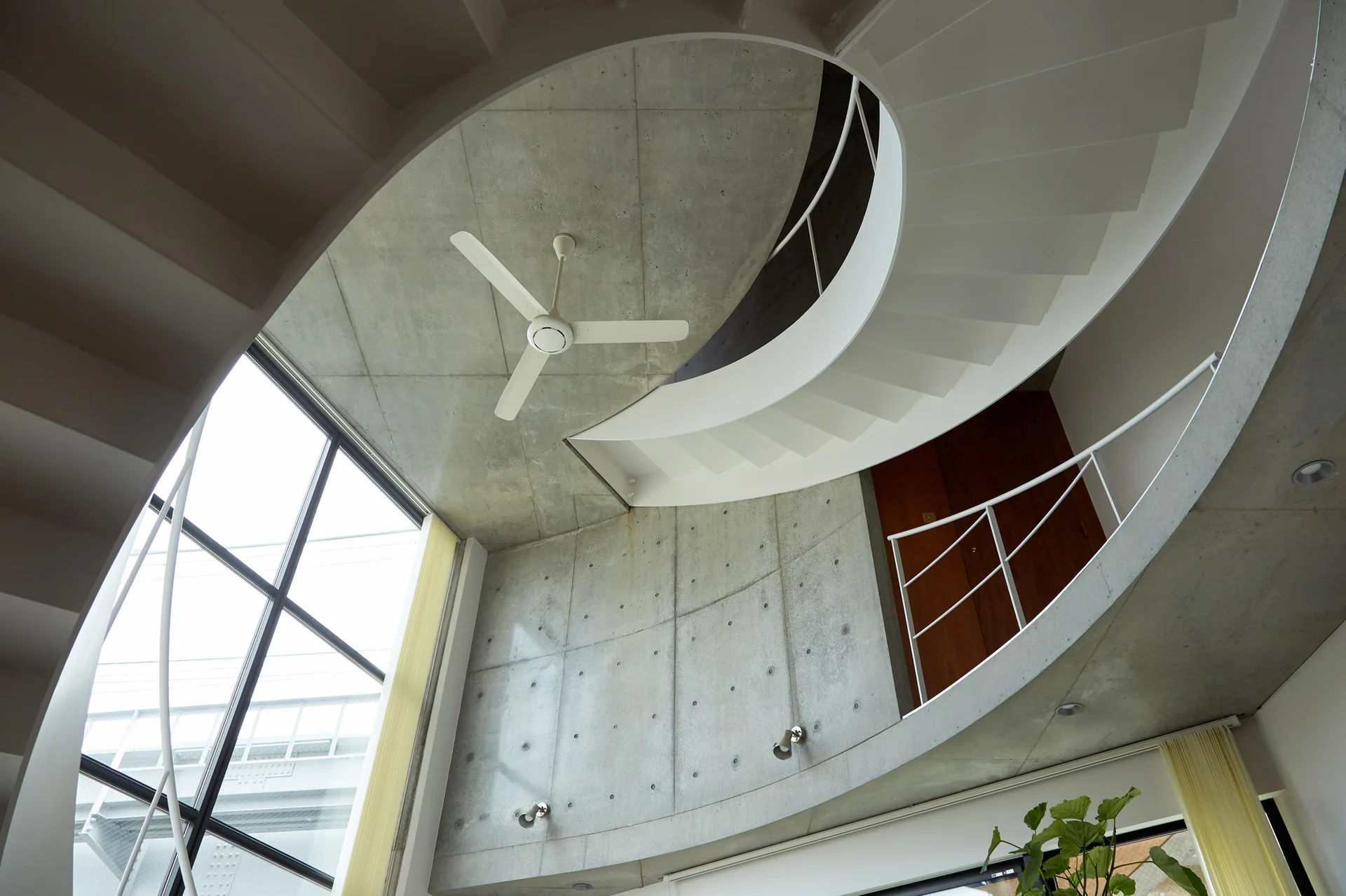A curved white spiral staircase winds up past an exposed raw concrete wall under a concrete ceiling with a white fan and a large gridded window.