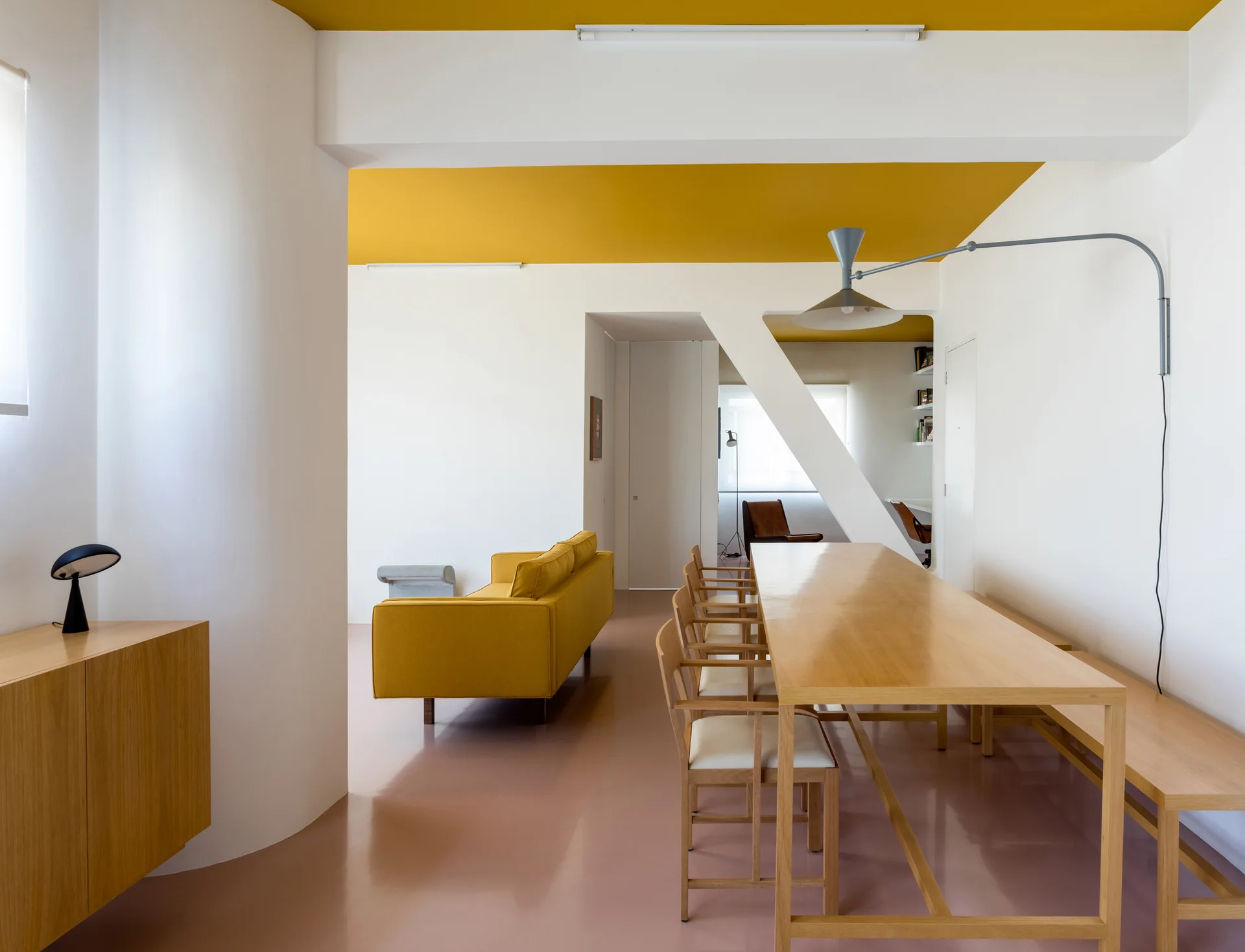 A modern open dining room with a mustard yellow ceiling, pale pink floor, long light wood dining table, and a yellow sofa.