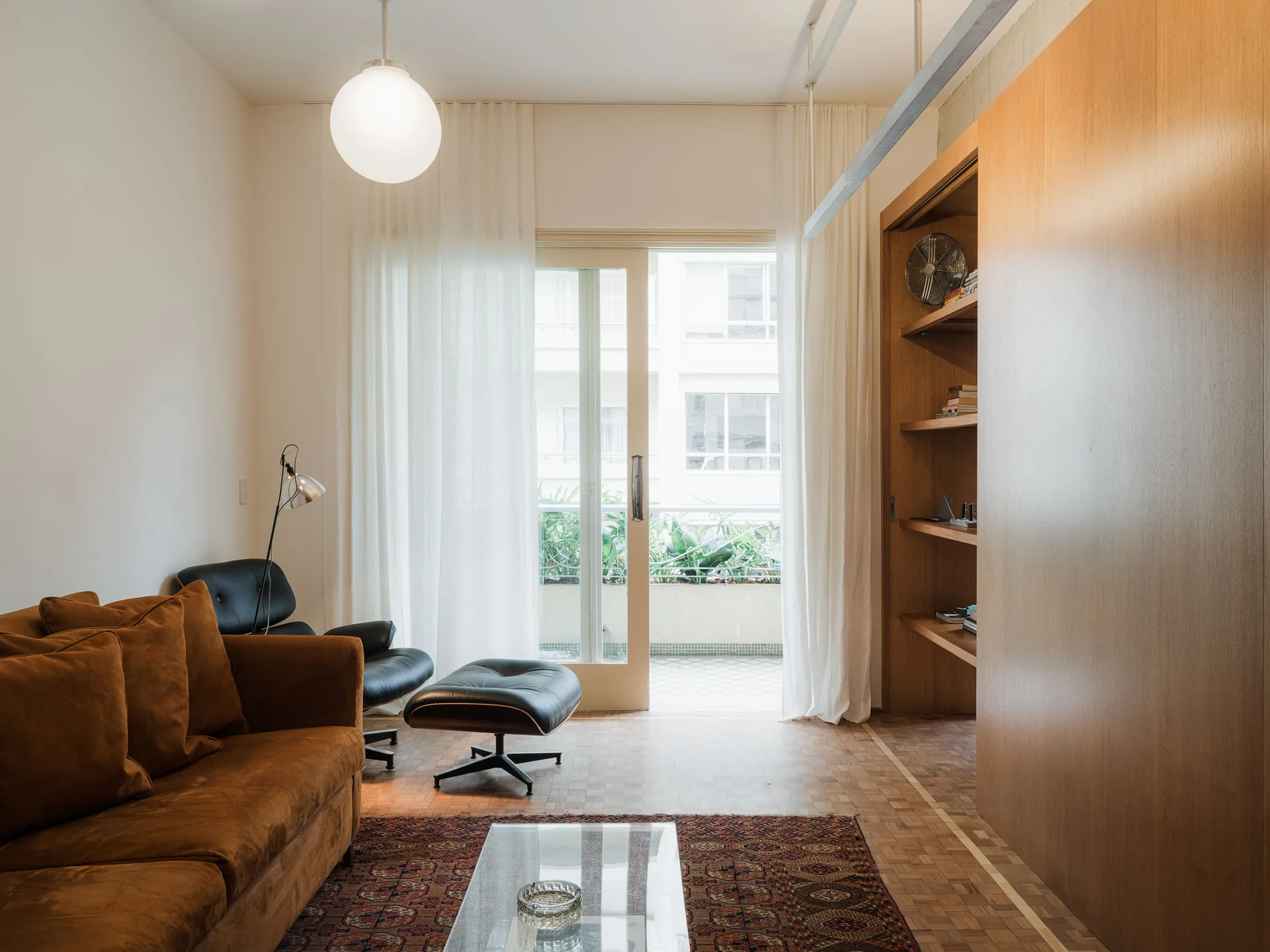 A living room with a brown velvet sofa, black leather lounge chair, parquet wood floor, and large wood-paneled wall unit leading to a balcony.