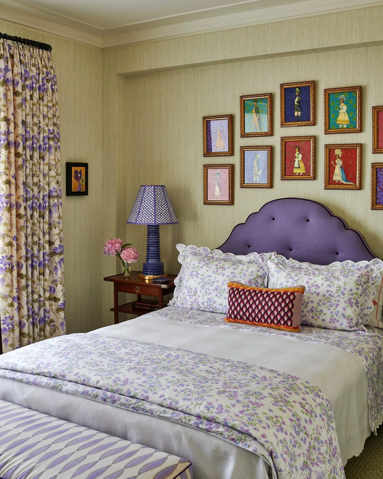 A bedroom features a purple scalloped headboard, floral bedding, and a gallery wall of small framed portraits on light green textured wallpaper.