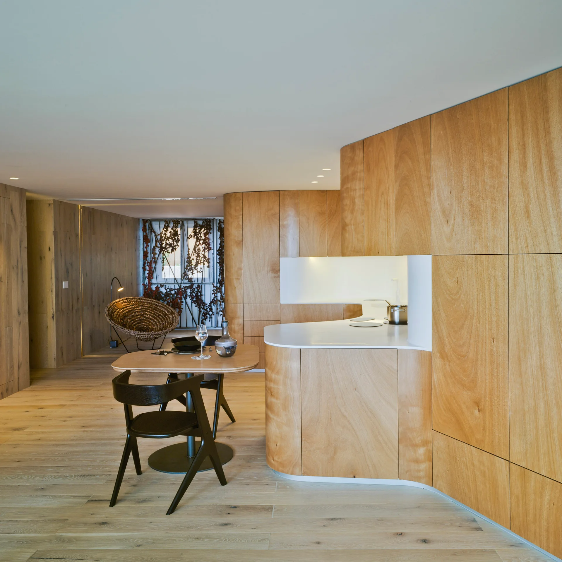 A minimalist kitchen features curved light wood cabinetry, white countertops, a small wooden dining table with black chairs, and wood flooring.