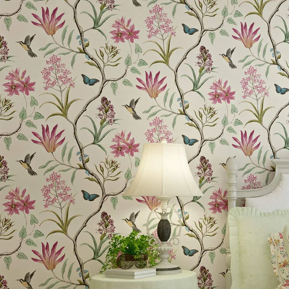 A bedroom corner features botanical wallpaper with pink flowers and birds behind a classic white table lamp and a green upholstered headboard.