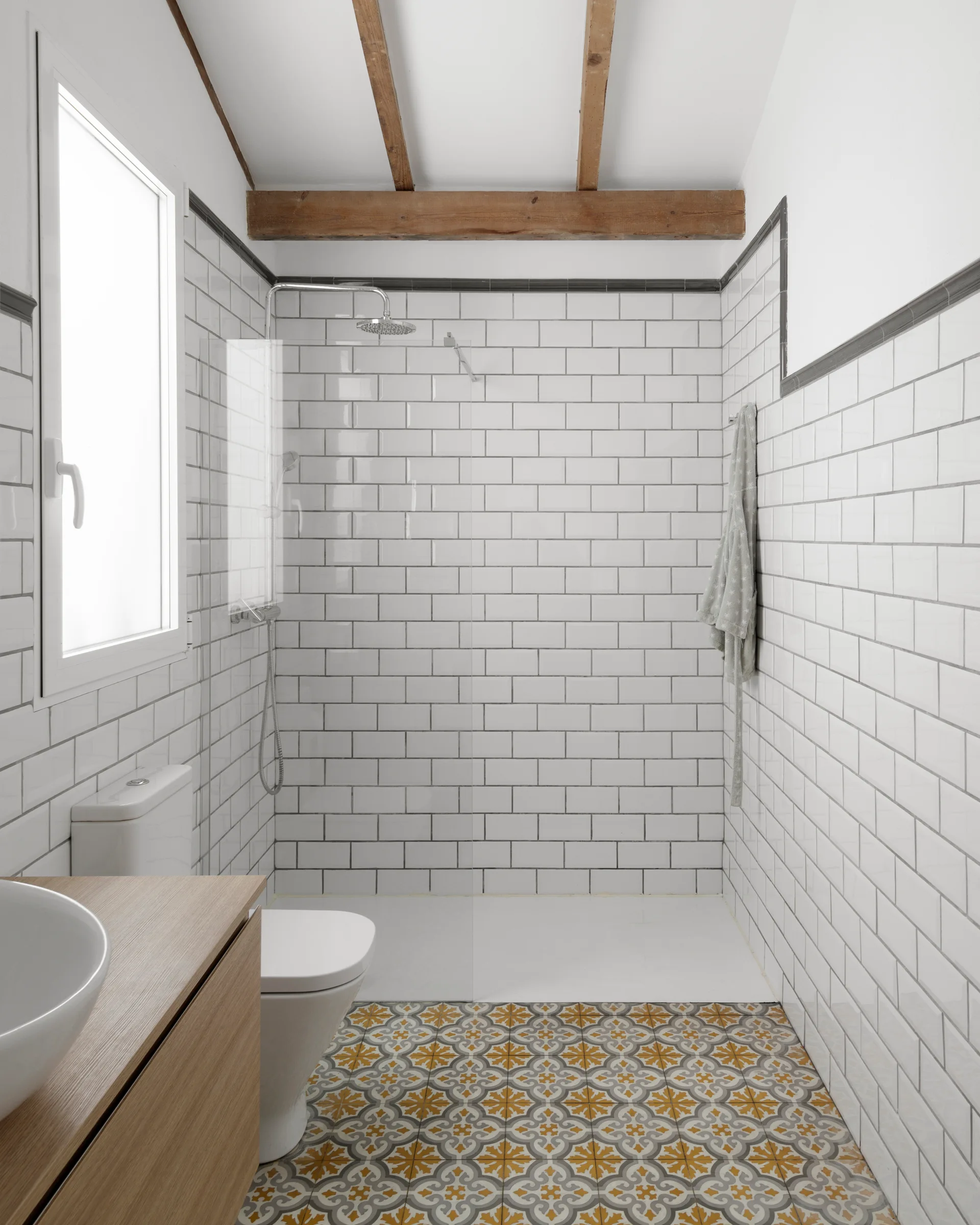 A bright bathroom features glossy white subway tile walls, patterned yellow and grey floor tiles, a wood vanity, and rustic wooden ceiling beams.