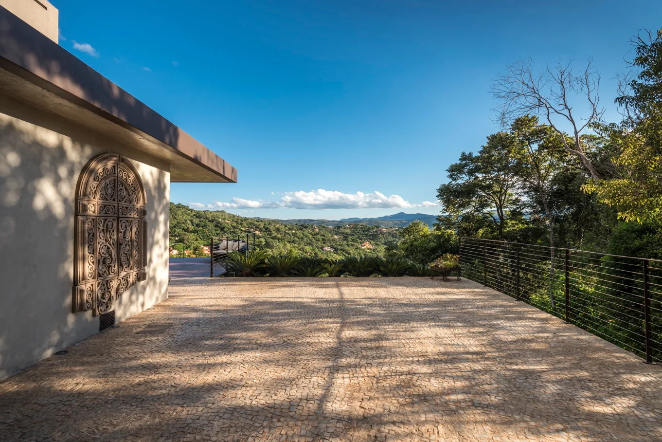 A wide cobblestone paved terrace features a flat plaster wall with a carved stone relief and a wire railing overlooking mountain views.