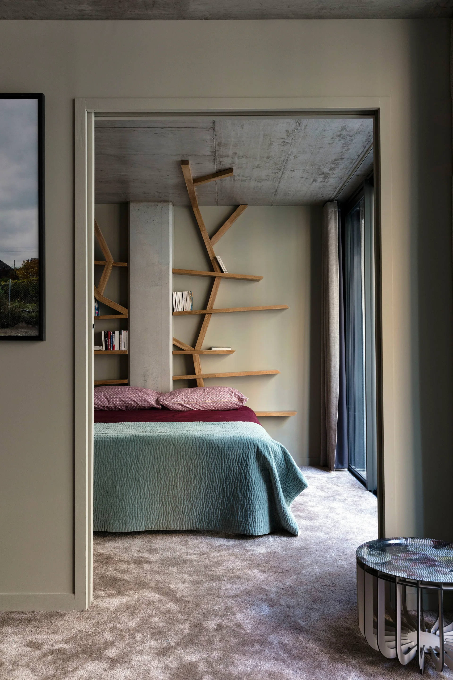 A modern bedroom viewed through a doorway features a bed with teal bedding, a branch-like wooden wall shelf, and an exposed concrete ceiling.
