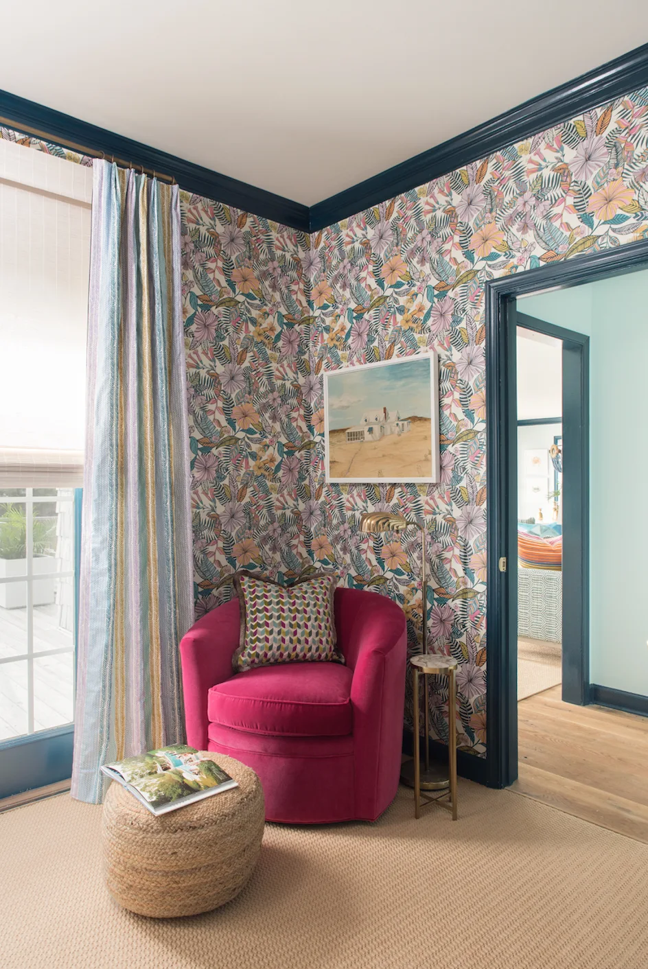 A pink armchair sits in a corner with floral wallpaper, a woven jute ottoman, striped curtains, and dark blue painted trim.