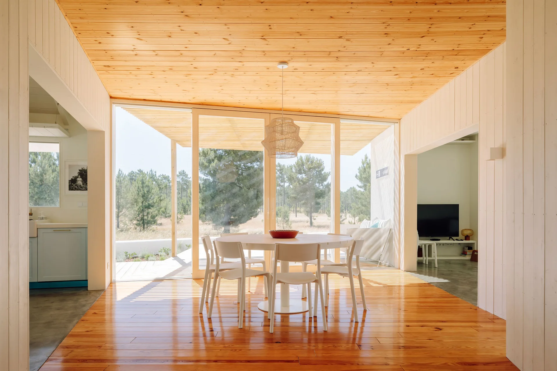 A minimalist dining room features a white round table, matching chairs, and a natural pine ceiling looking out to a forest through large glass doors.
