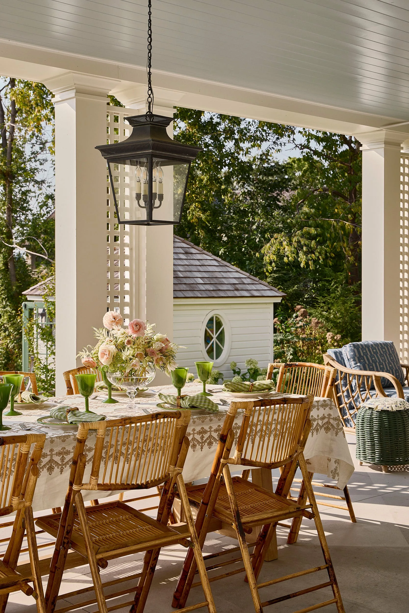 A covered porch dining table is set with a patterned tablecloth, green glassware, and a floral centerpiece, surrounded by bamboo folding chairs.