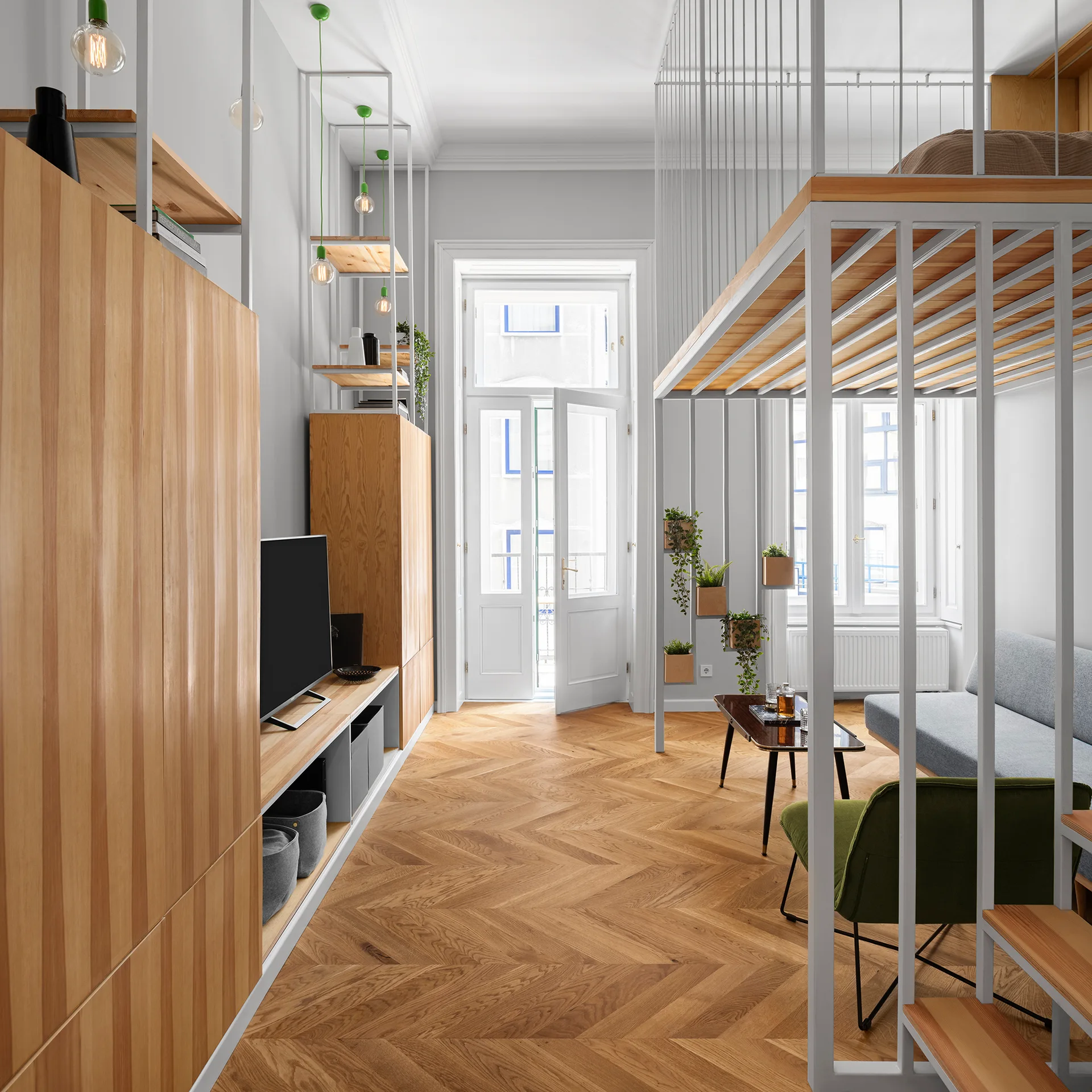 A contemporary studio apartment featuring a metal loft bed frame, built-in wooden cabinets, a grey sofa, and herringbone patterned wood floors.