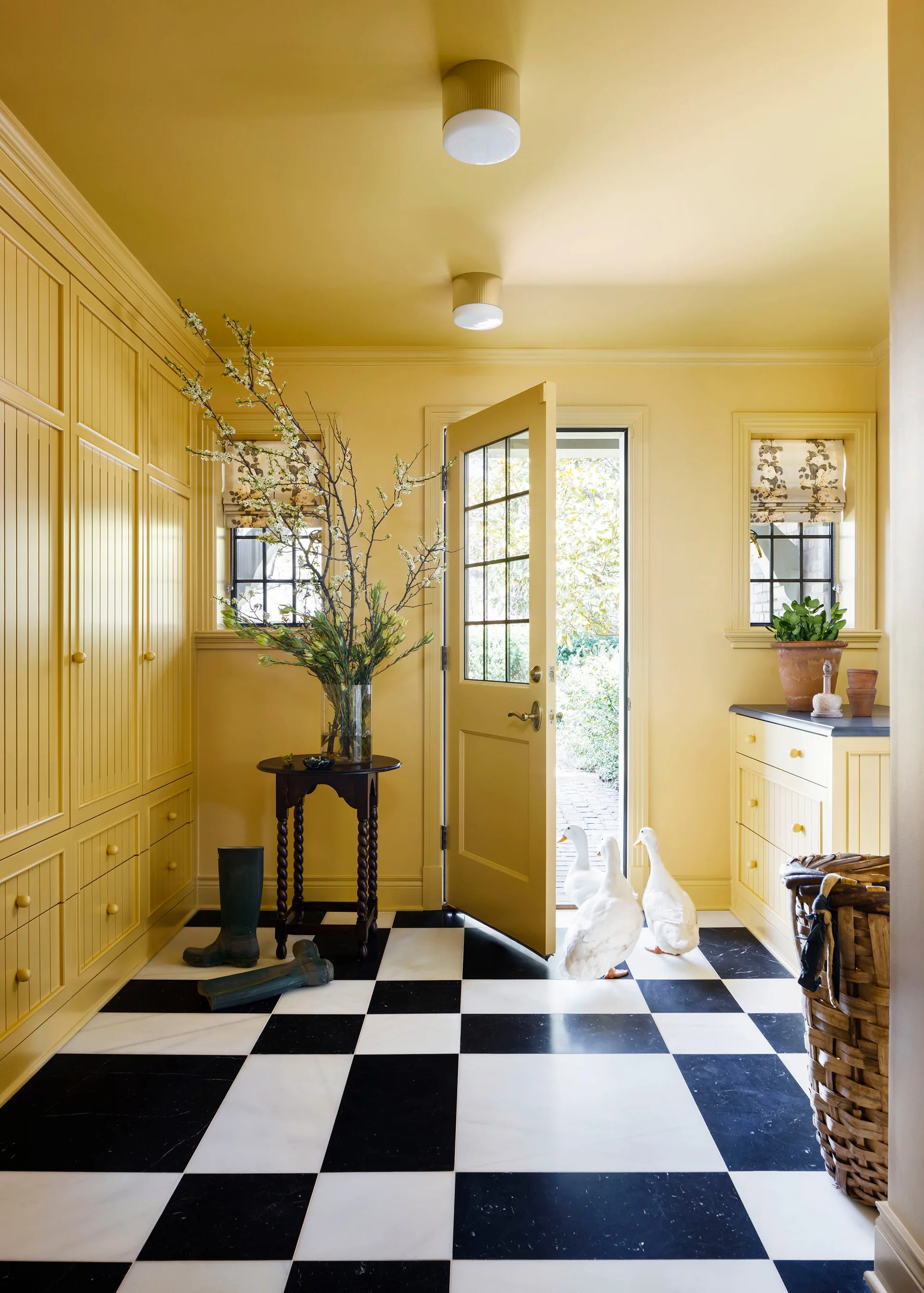 Three white ducks enter a yellow mudroom featuring built-in cabinetry, a dark wood table, and a black-and-white checkerboard tile floor.