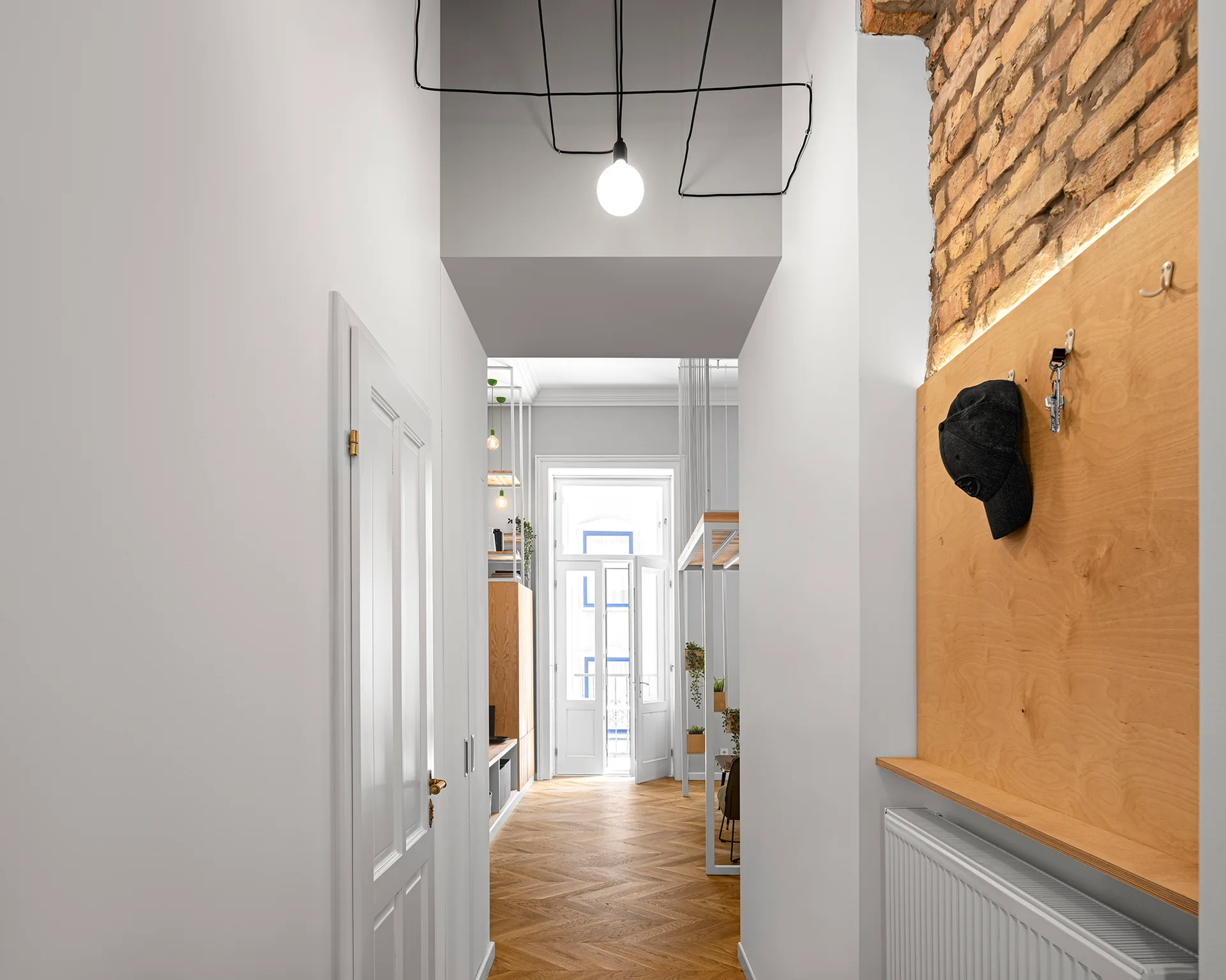 A bright hallway featuring white walls, chevron wood flooring, and a plywood wall panel topped with exposed brick.