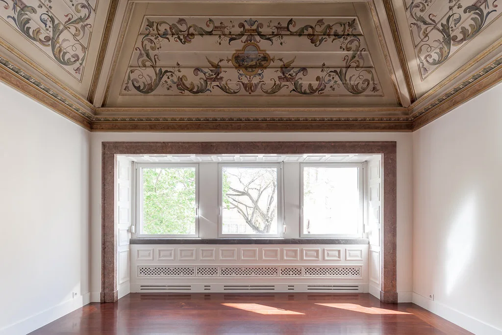 An empty room features a polished wood floor, white walls, a triple window framed in reddish stone, and a highly decorative painted ceiling.