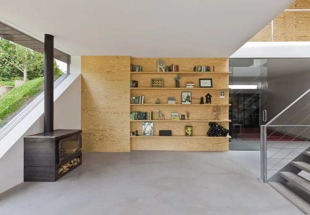 A minimalist living room featuring a custom plywood wall with built-in shelves, a black wood stove, and a polished concrete floor.