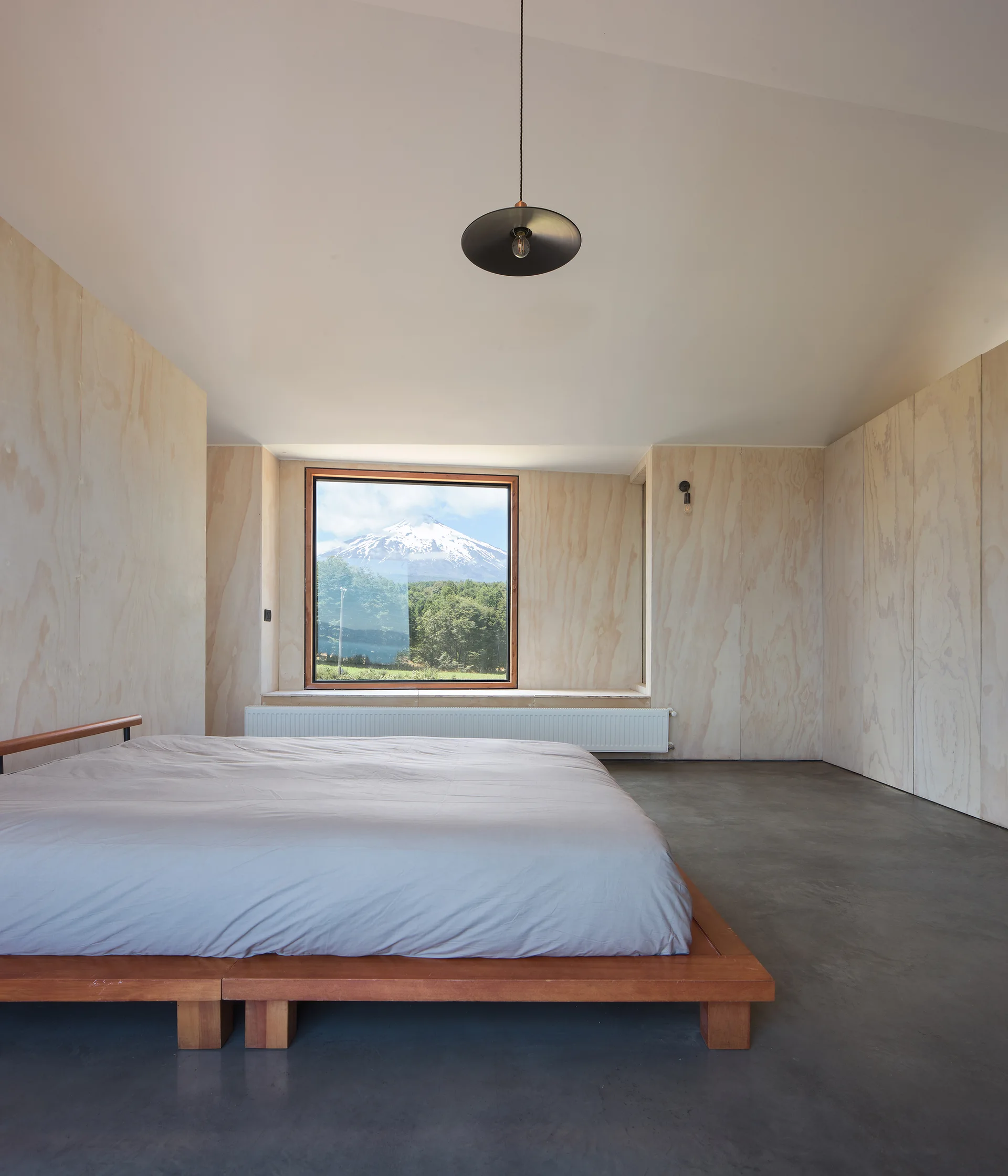 A minimalist bedroom featuring natural plywood paneled walls, a low wooden platform bed, and a square window framing a snow-capped mountain.