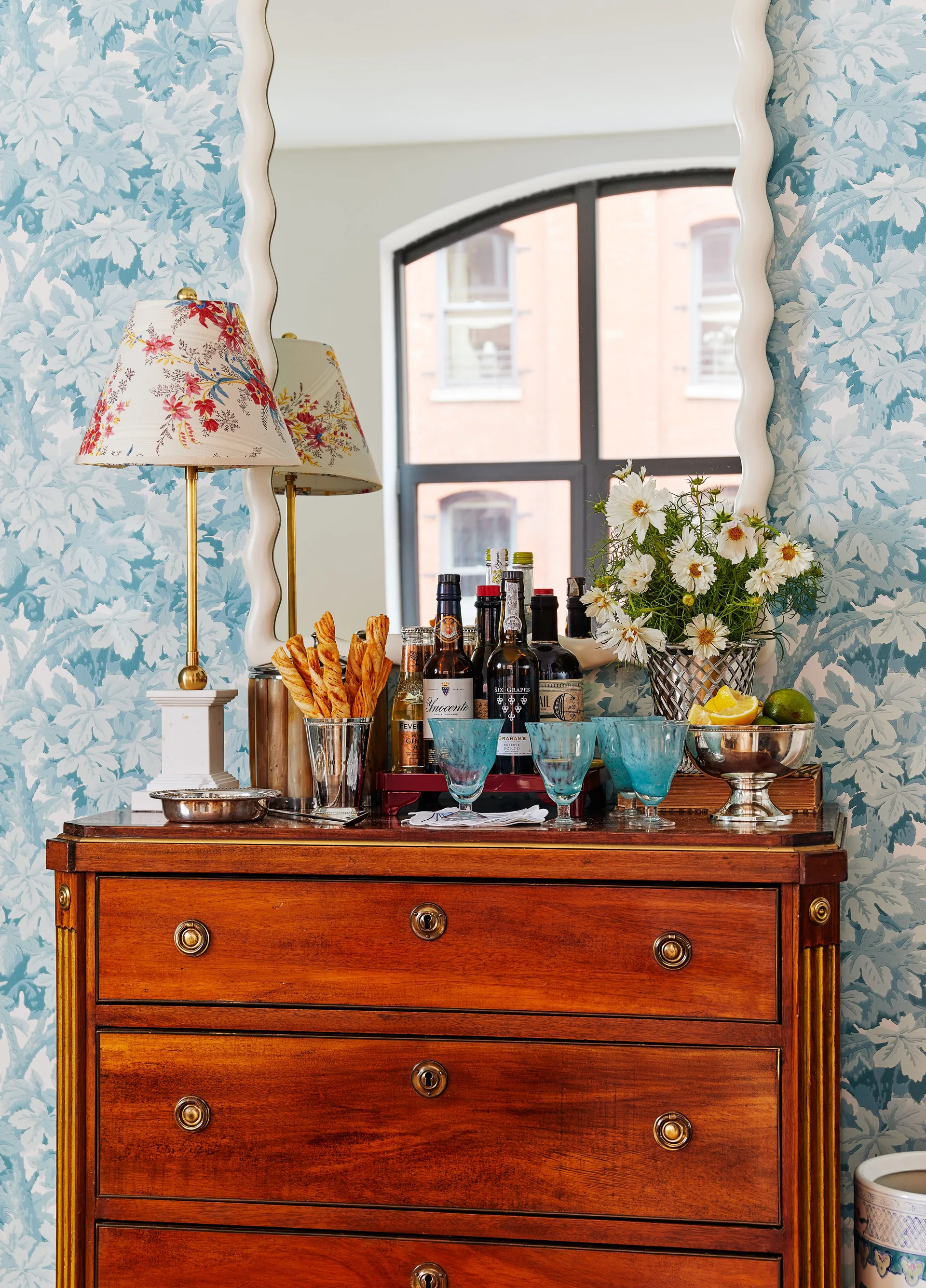A wooden chest of drawers set up as a bar with glassware and bottles against blue floral wallpaper beneath a white wavy mirror.