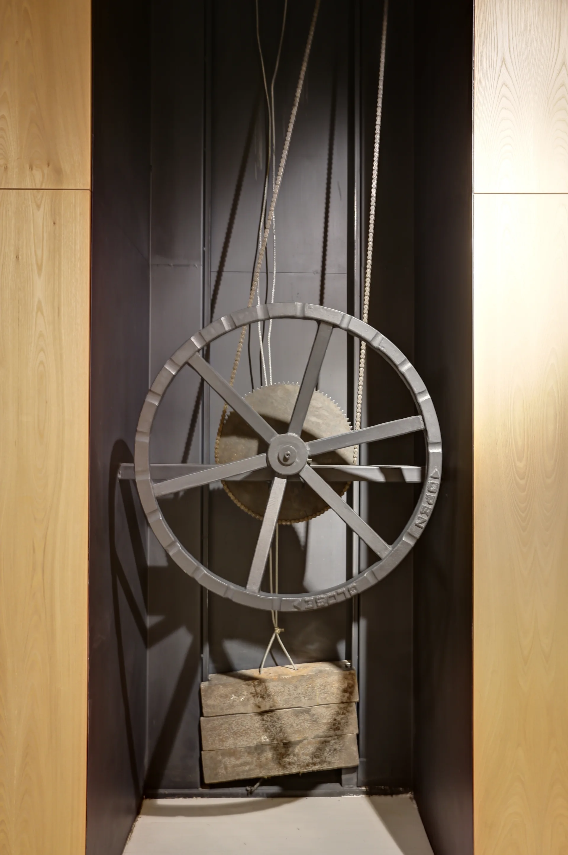 A mechanical metal pulley system with a large wheel and counterweight is recessed in a dark niche flanked by light wood wall panels.