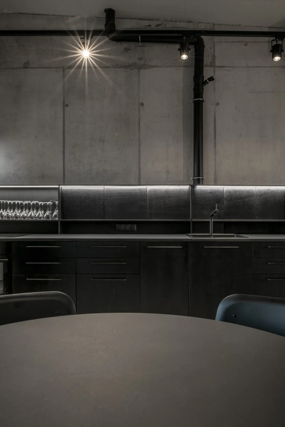 An industrial kitchen features matte black cabinetry, an integrated sink, a raw concrete wall, and overhead track lighting.