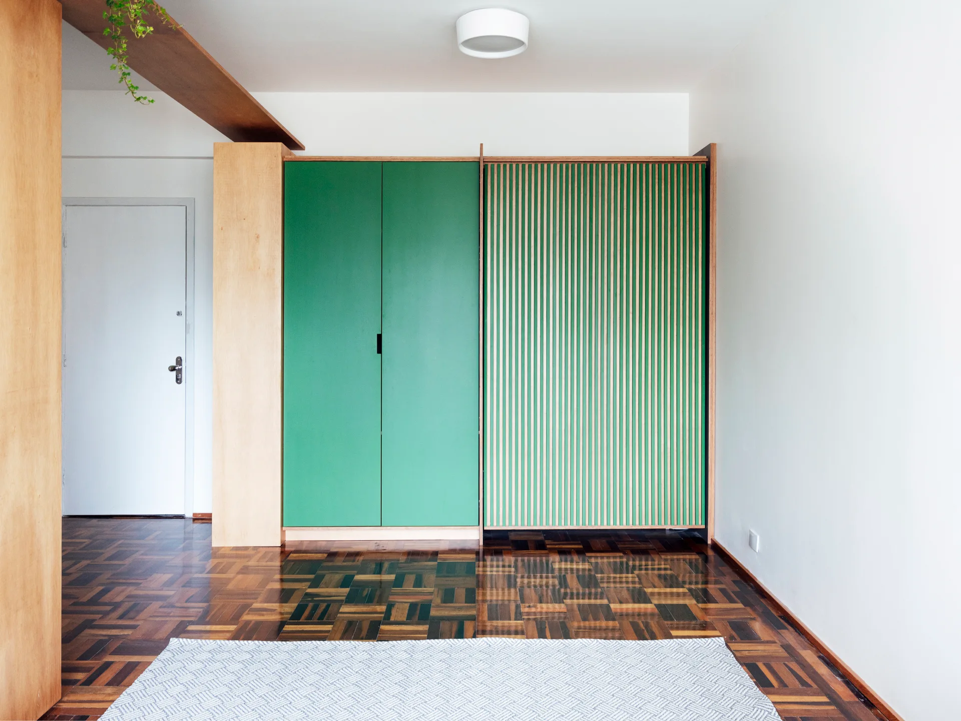 A custom floor-to-ceiling wardrobe with solid green and slatted green doors stands on patterned parquet wood flooring in a minimalist room.