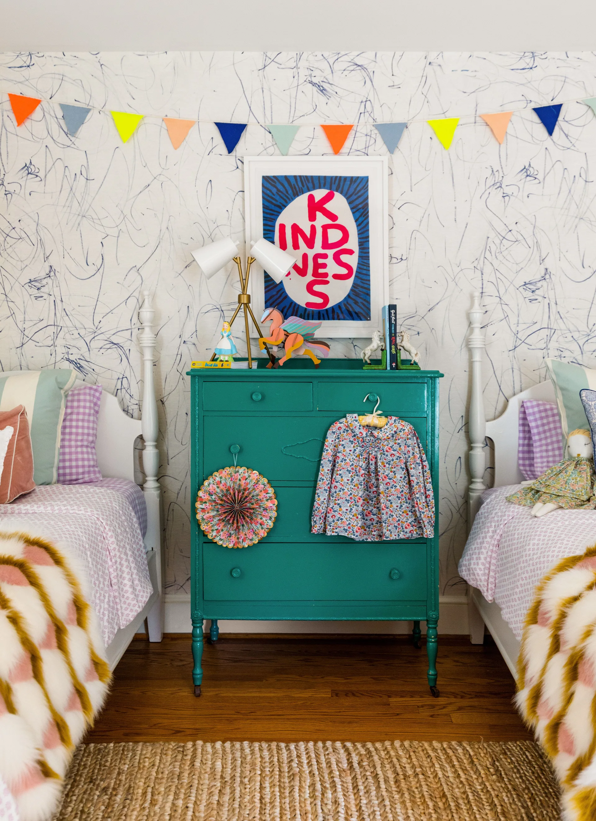 Two white twin beds flank a bright teal wooden dresser against scribble-patterned wallpaper in a playful children's bedroom.