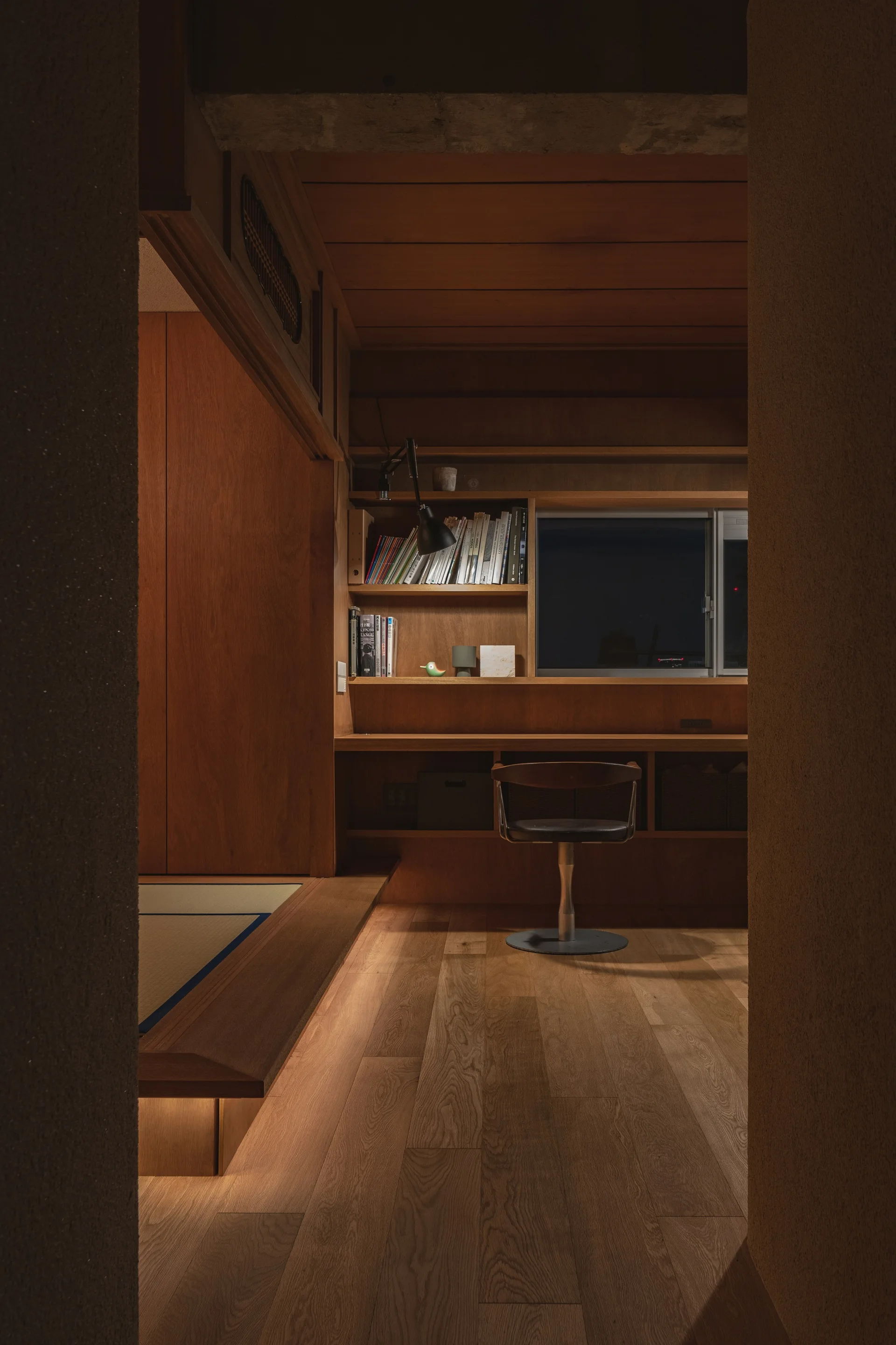 A dark-wood paneled home office features a built-in desk with a black task lamp, a swivel chair, and a raised tatami platform.