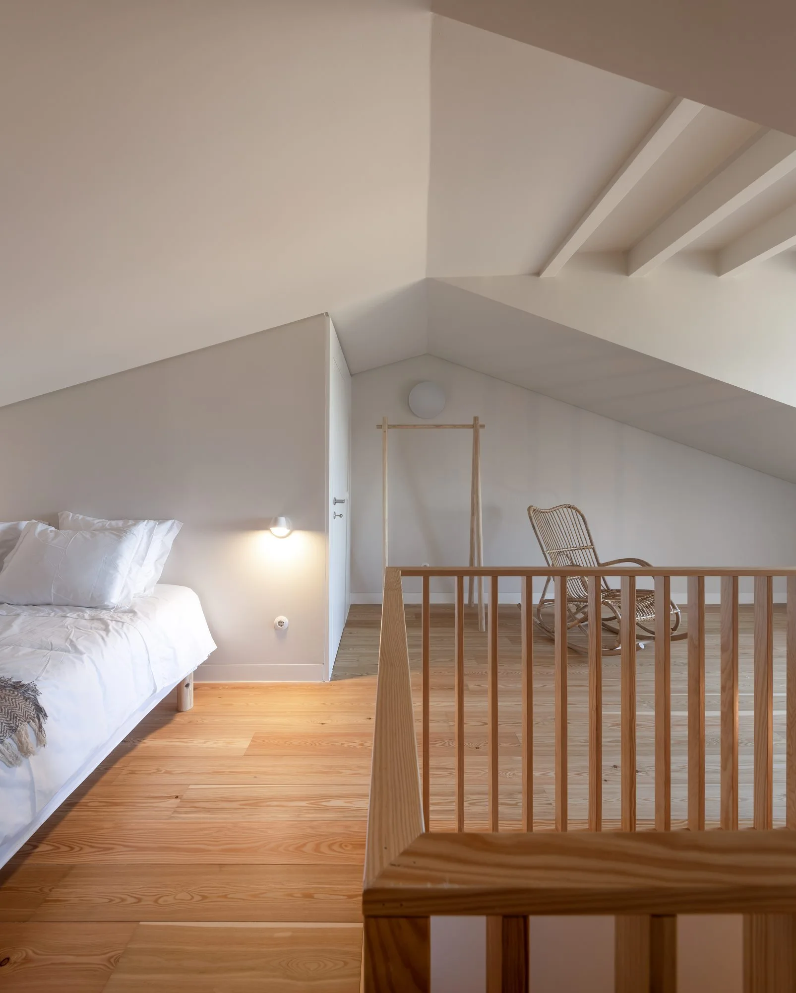 An attic bedroom with a low bed, a wicker rocking chair, a wooden staircase railing, and sloped ceilings over light wood flooring.