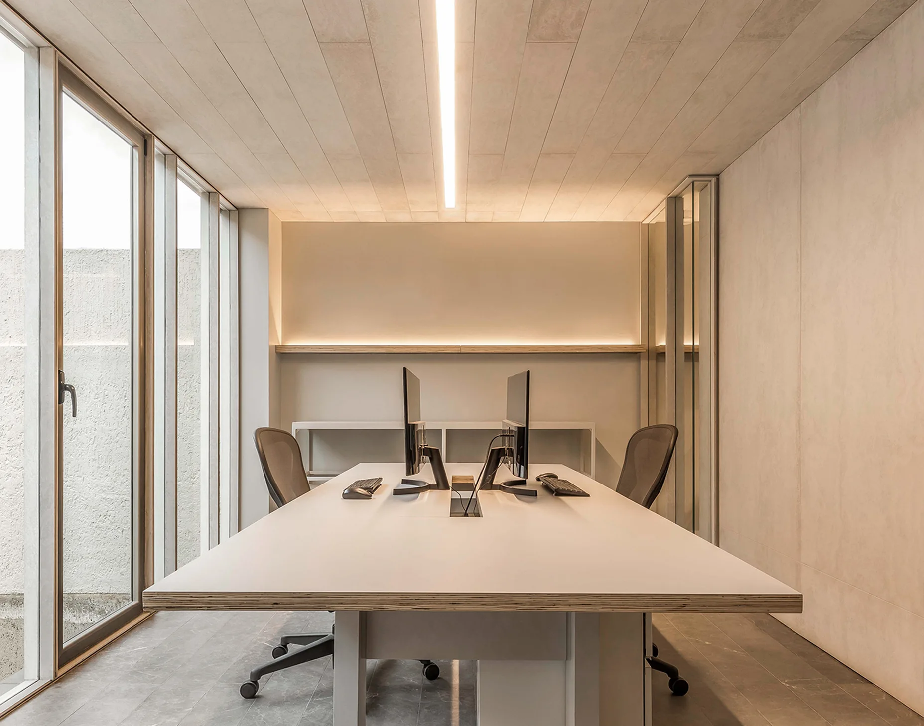 A contemporary minimalist home office featuring a large white desk with two monitors under a wood plank ceiling with a linear light fixture.
