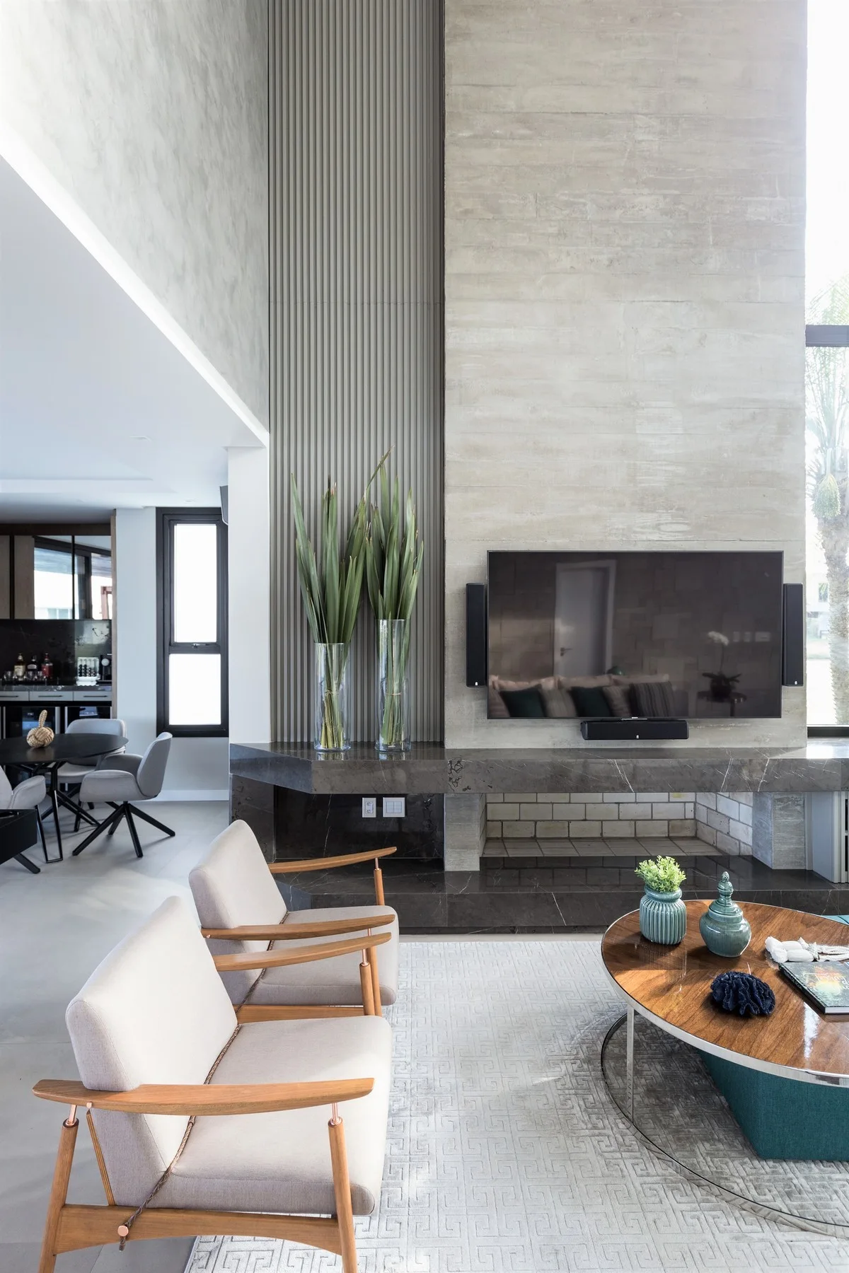A modern living room features two light grey armchairs facing a round wooden coffee table, set before a tall concrete fireplace with ribbed wall paneling.