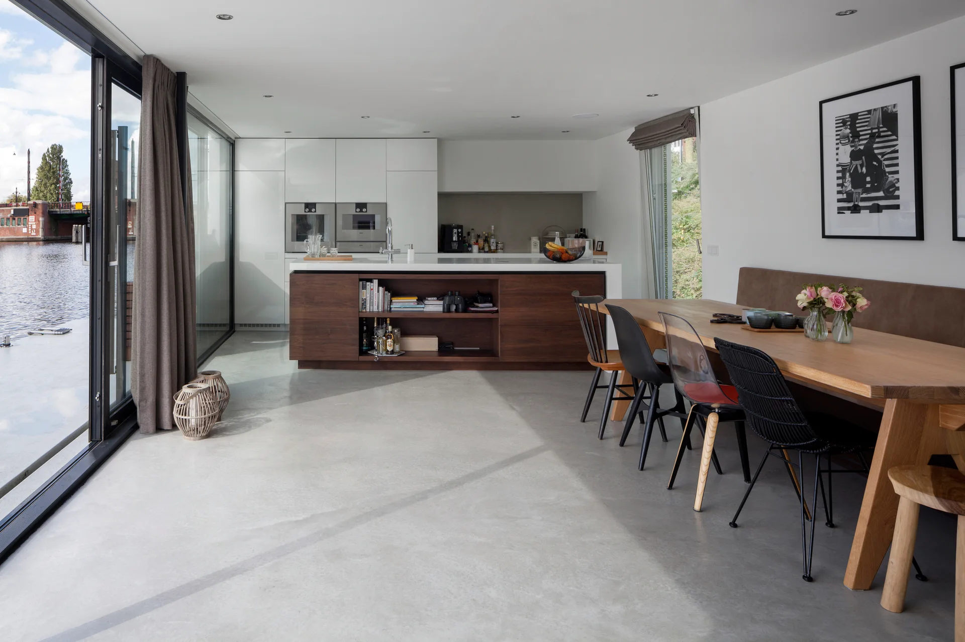 A modern open-concept kitchen and dining area features a wood island, eclectic dining chairs at a wooden table, and sliding glass doors overlooking water.