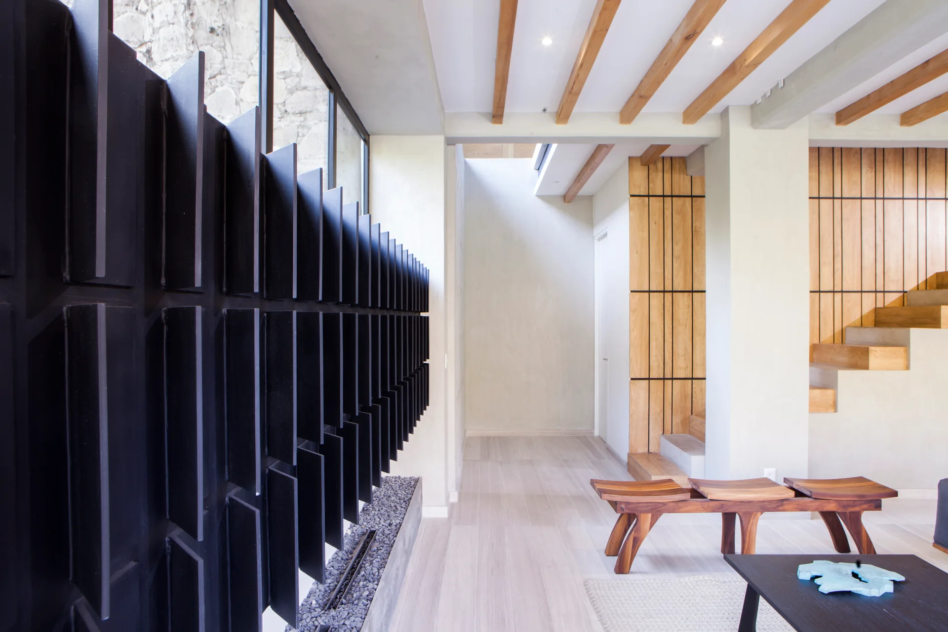 A contemporary living space featuring a sculptural black metal divider, exposed wooden ceiling beams, and a curved wood bench.
