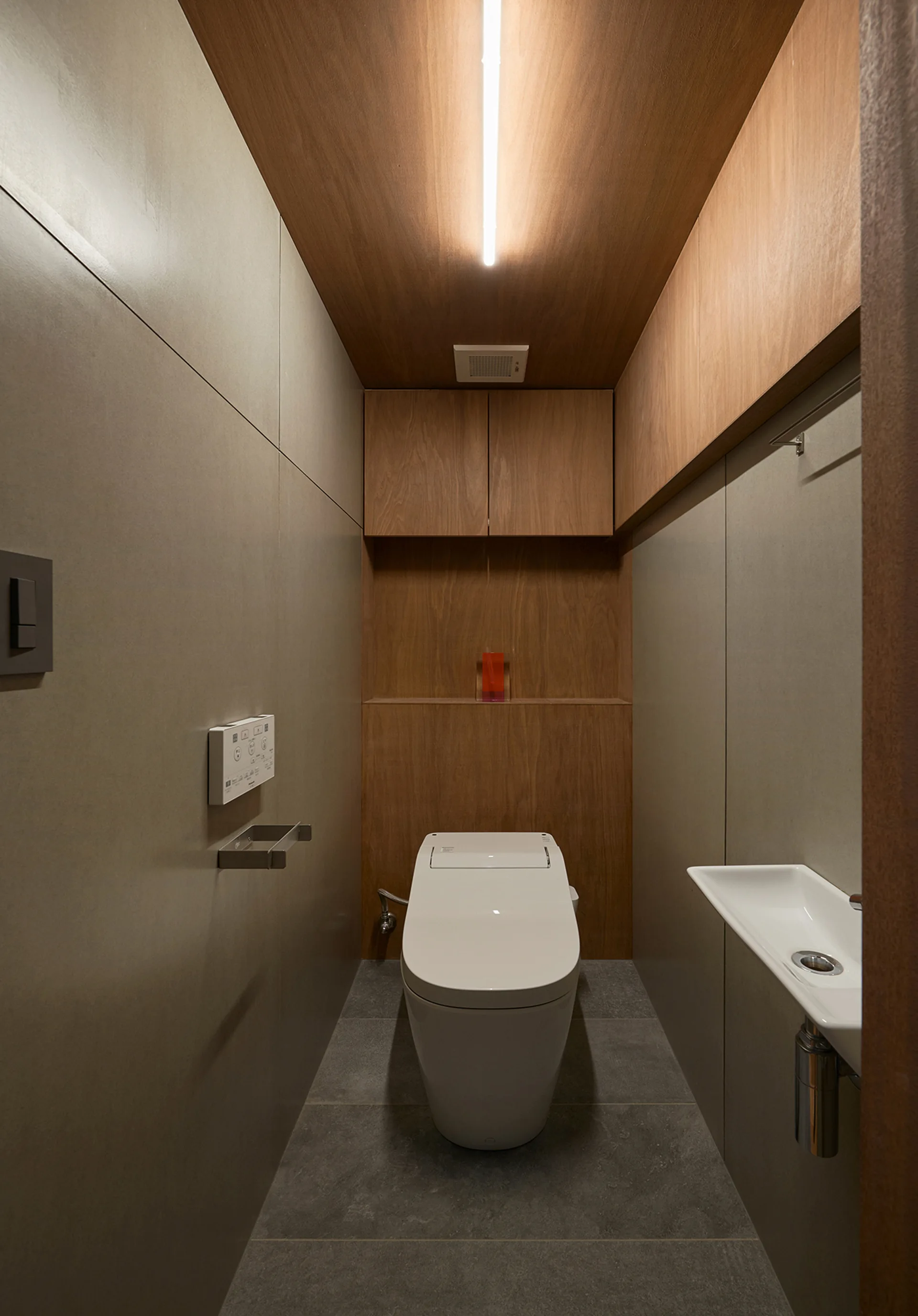 A narrow modern powder room features grey paneled walls, a wood ceiling, a built-in wooden cabinet, a white toilet, and a tiny wall-mounted sink.